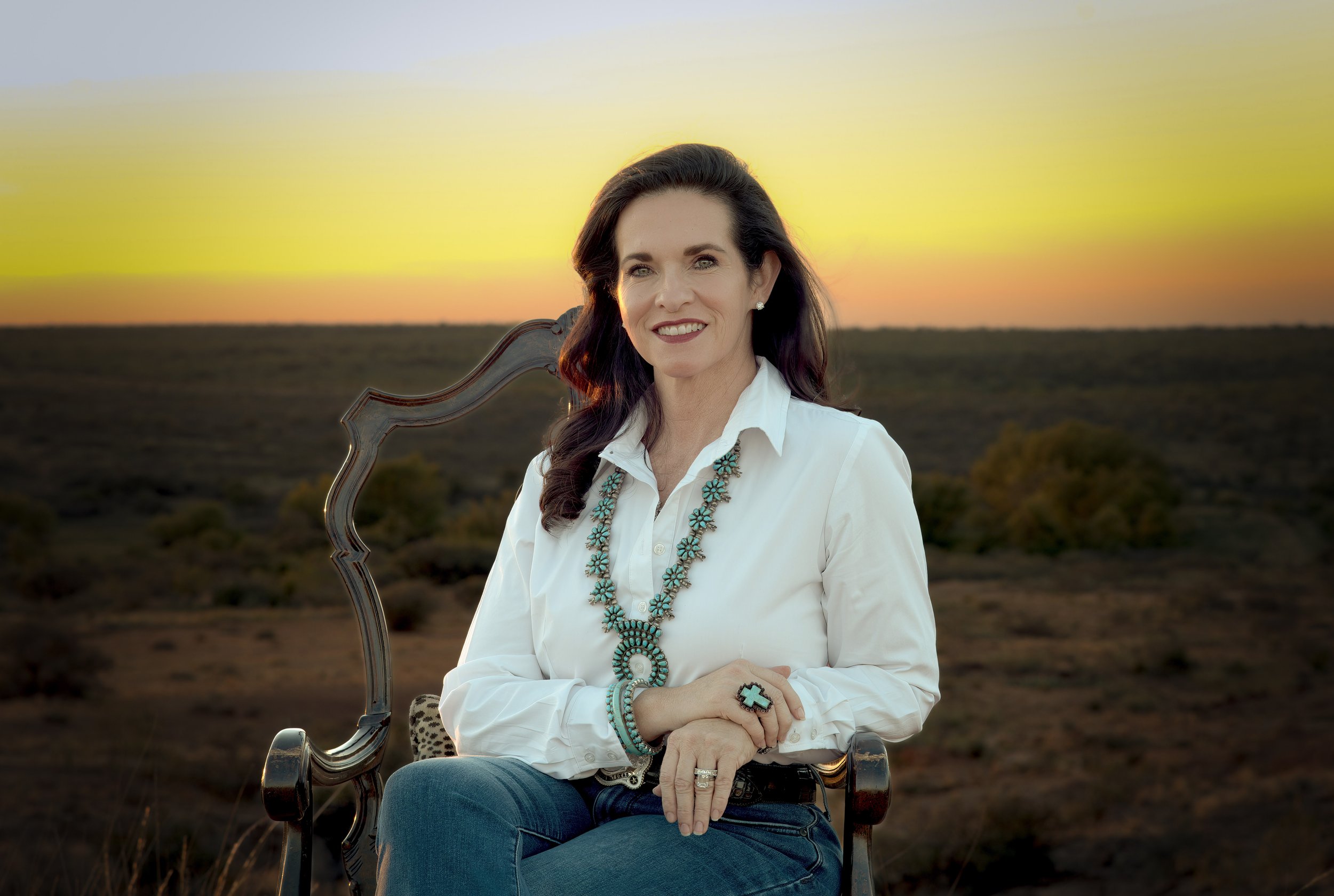 Smiling woman in a white shirt and turquoise jewelry seated in a wooden chair at sunset in an open desert landscape