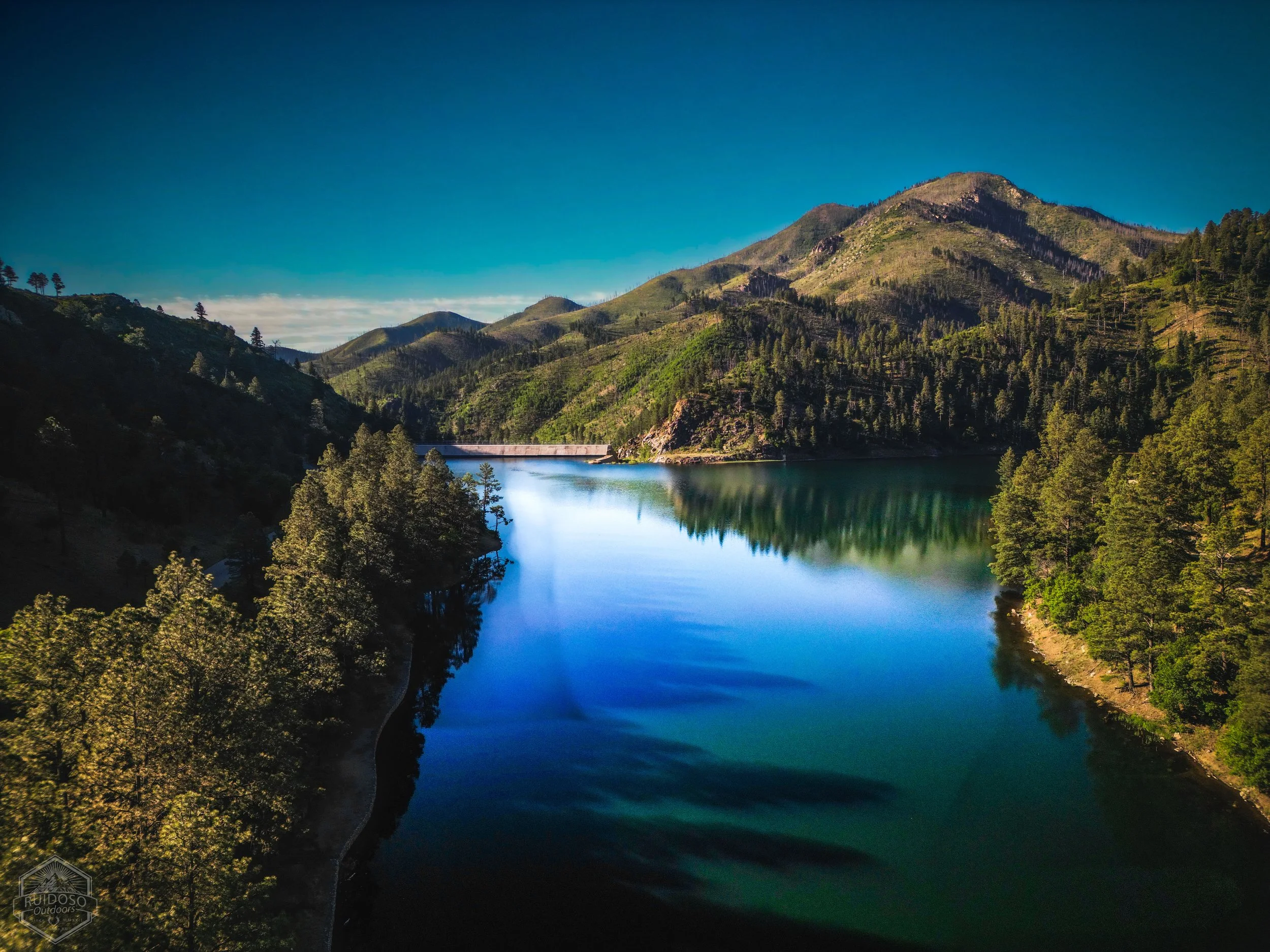 Aerial view of a forested mountain lake and dam beneath a clear blue sky