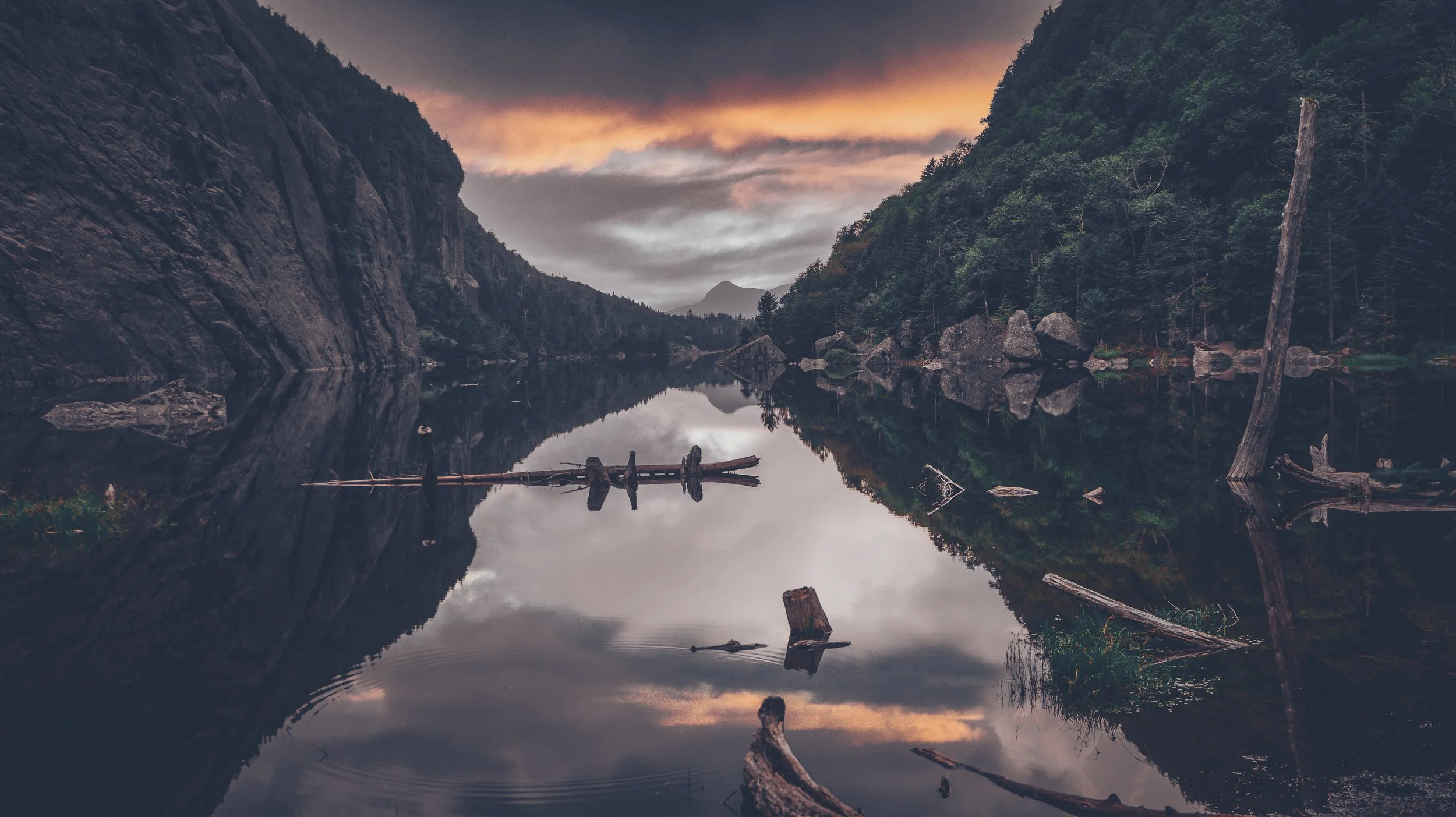 Serene mountain lake reflecting forested cliffs, distant peaks, fallen logs, and a dramatic sunset sky