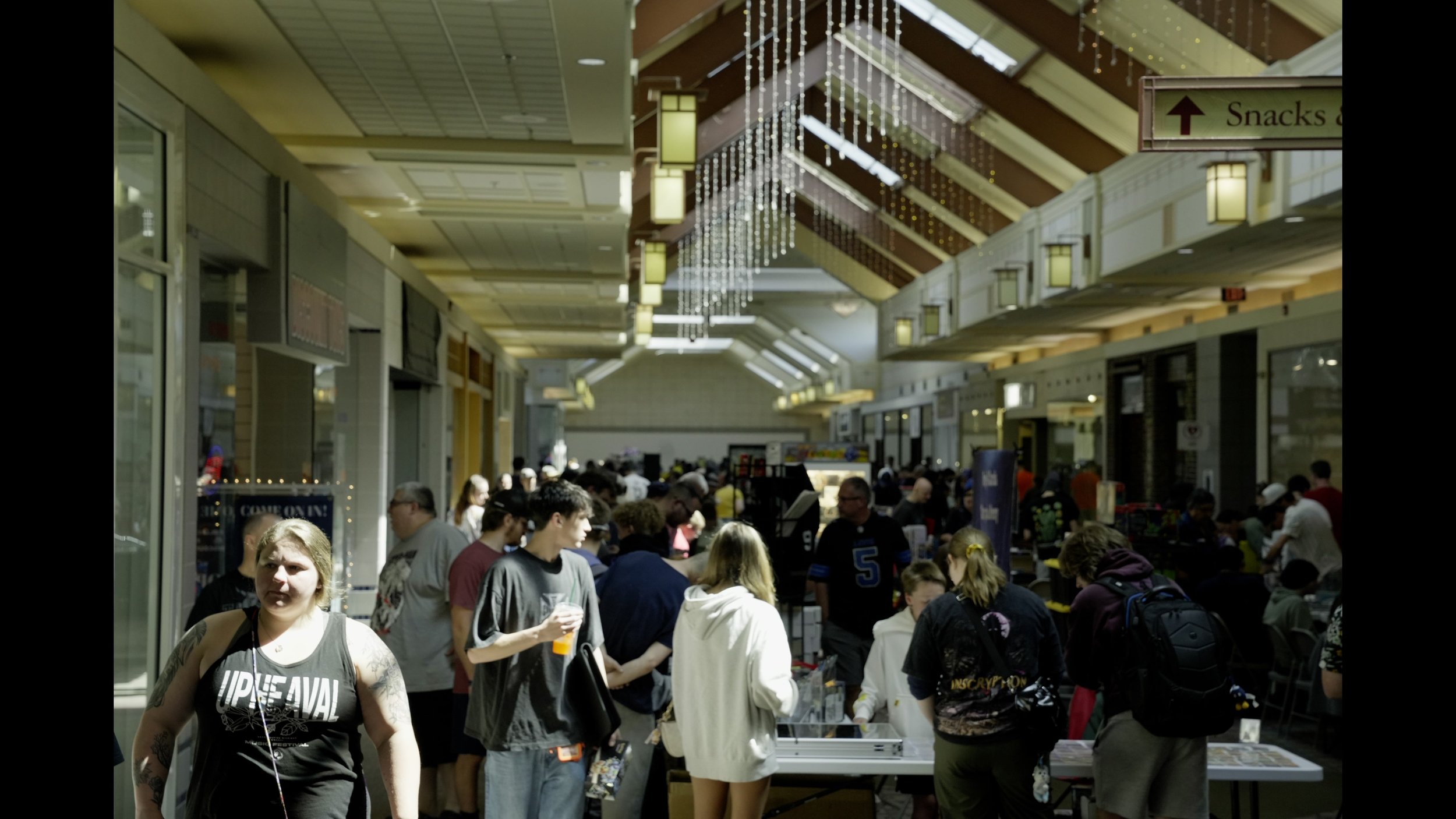 Crowded shopping mall atrium with shoppers, storefronts, hanging lights, and a sign pointing to snacks.