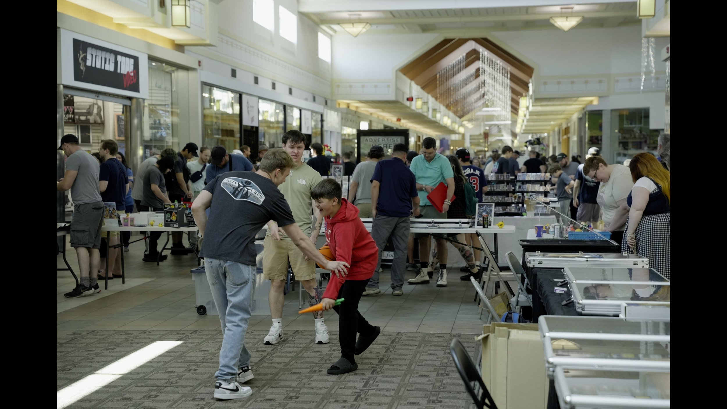 Crowded indoor marketplace with vendors selling collectibles and people browsing tables in a shopping mall corridor