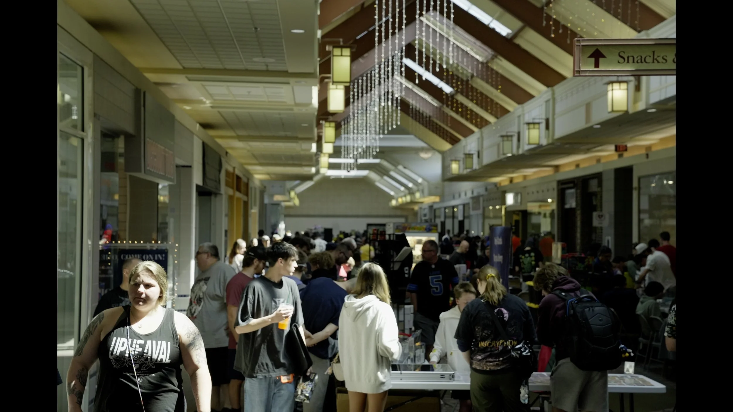 Crowded shopping mall corridor with shoppers, vendor tables, skylights, and a sign pointing to snacks.