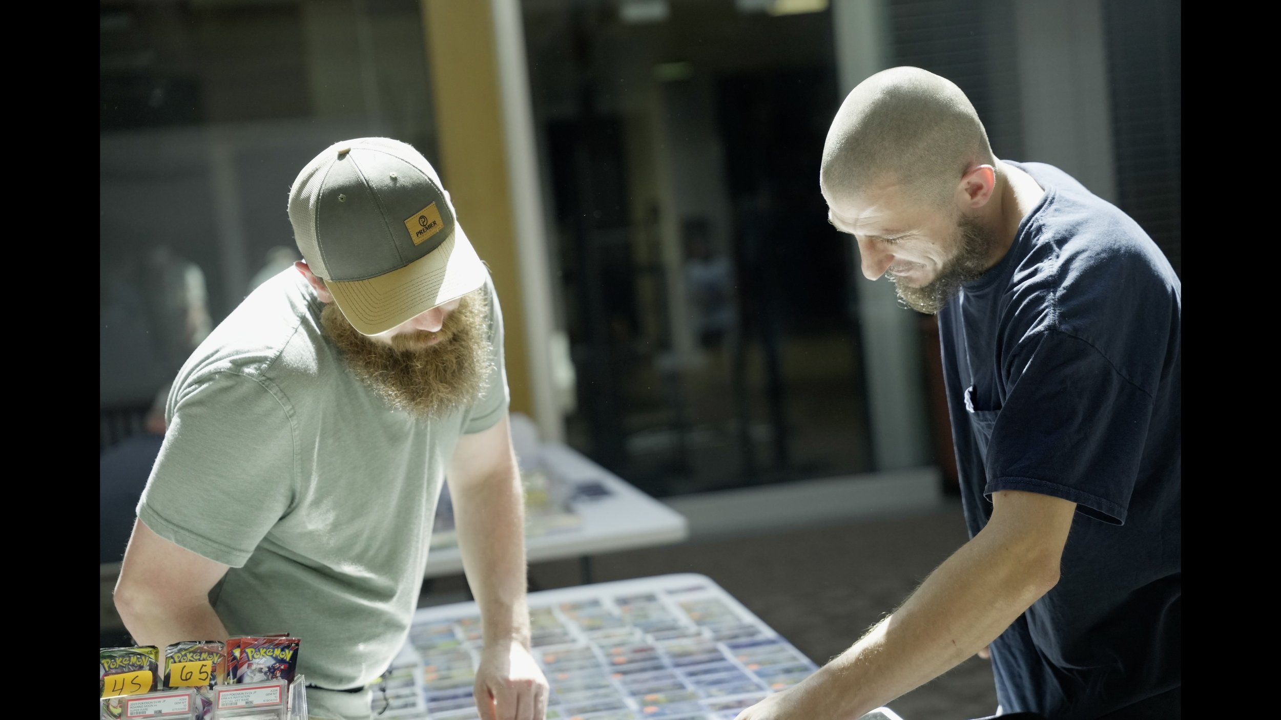 Two bearded men examine Pokémon trading cards at a card market table.