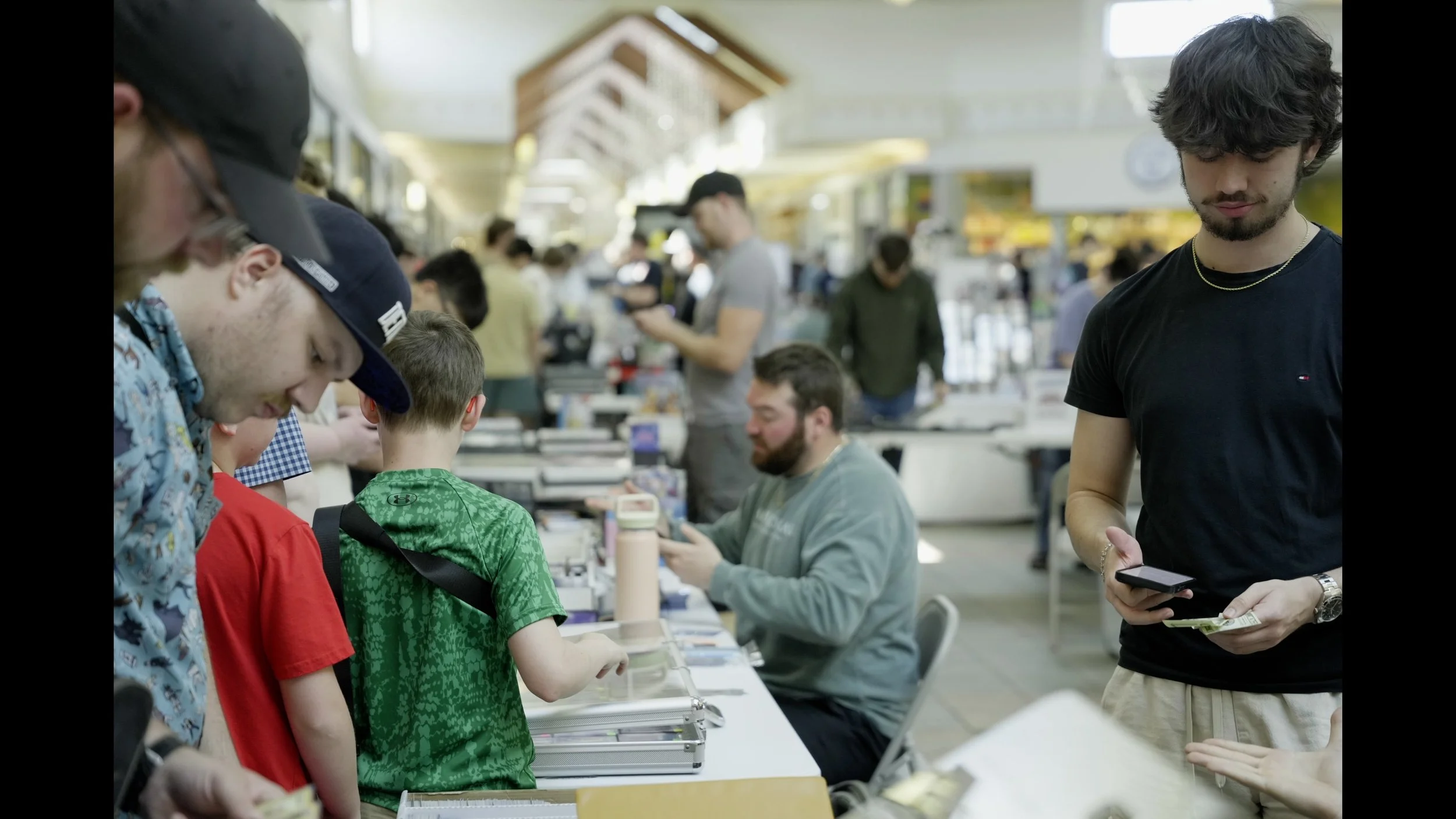 Crowded indoor event with attendees browsing tables; a young man in a black shirt checks his phone in the foreground.