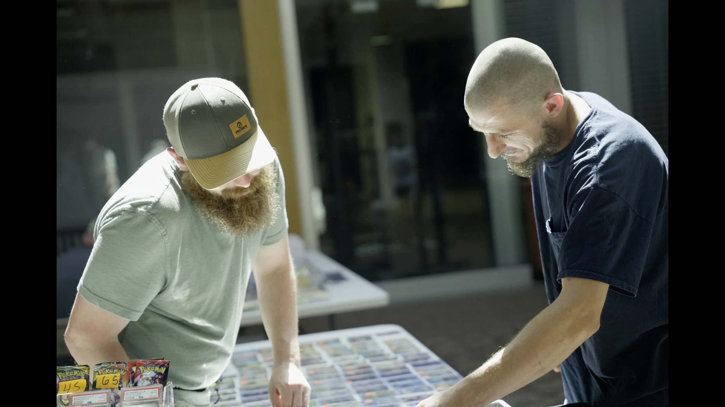 Two bearded men examine Pokémon trading cards at an outdoor vendor table.