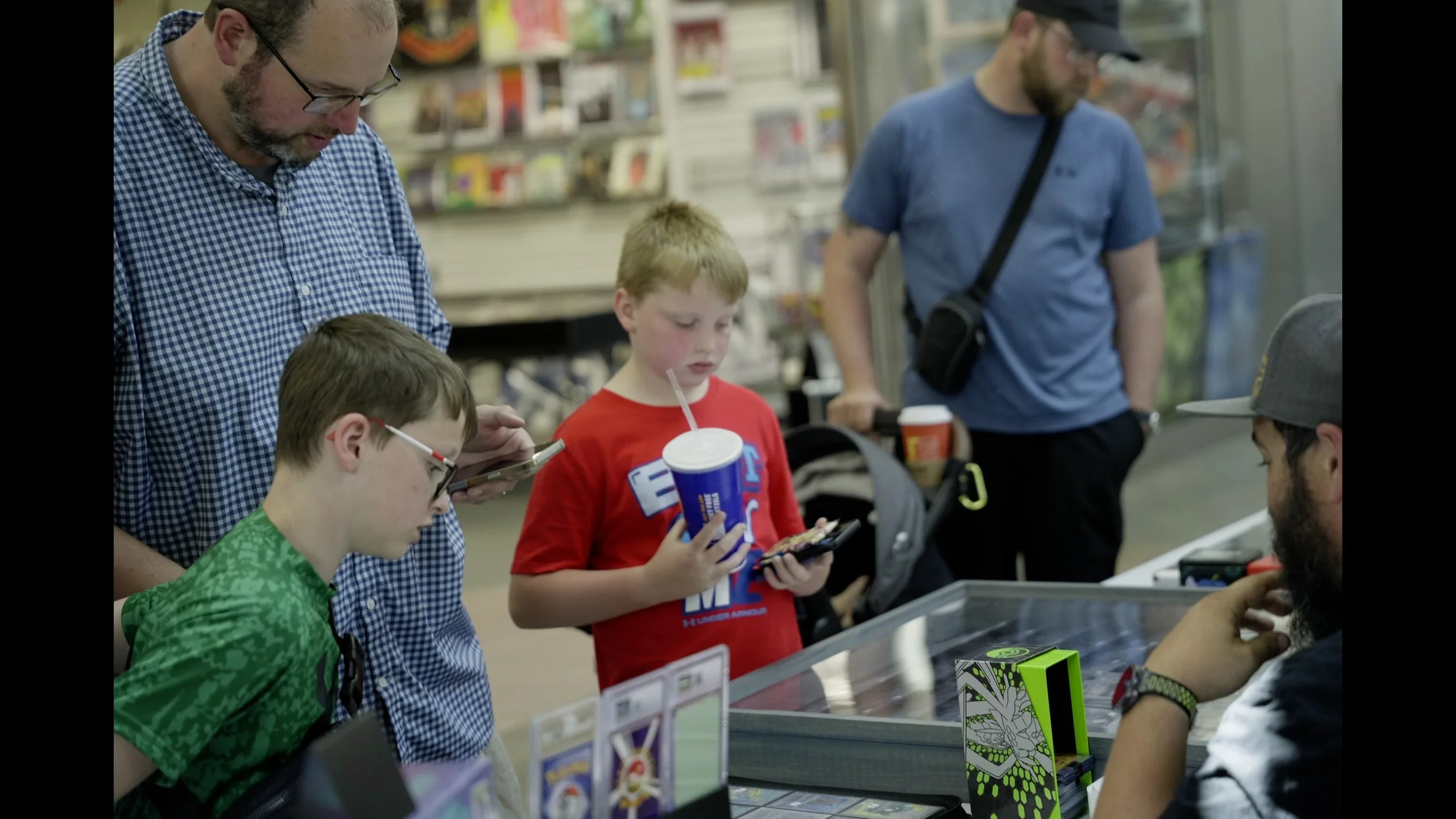 Family browsing collectibles at a store counter, with two boys holding drinks and trading cards.