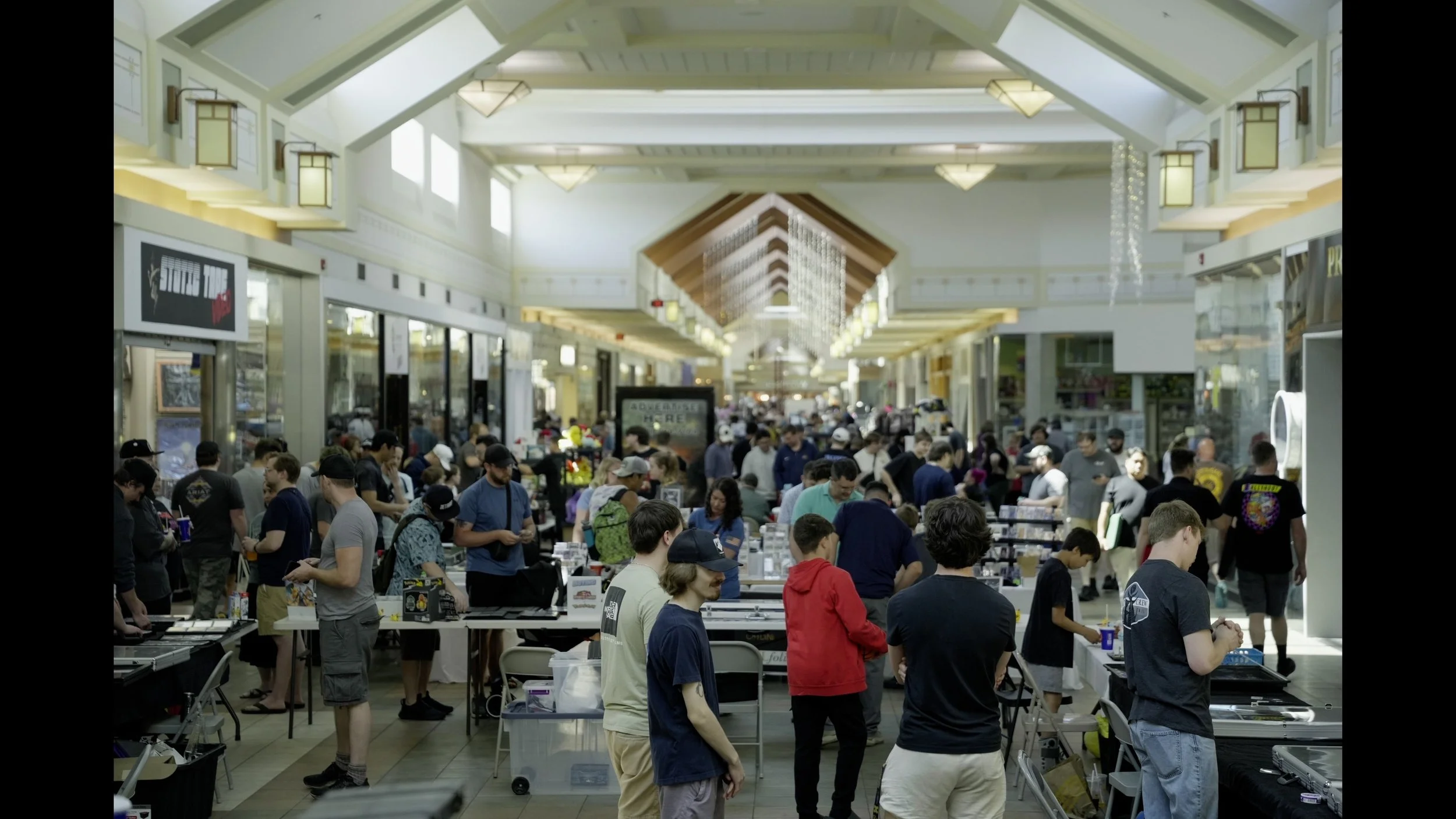 Crowded indoor convention hall with vendors, merchandise tables, and shoppers beneath ornate Art Deco lighting