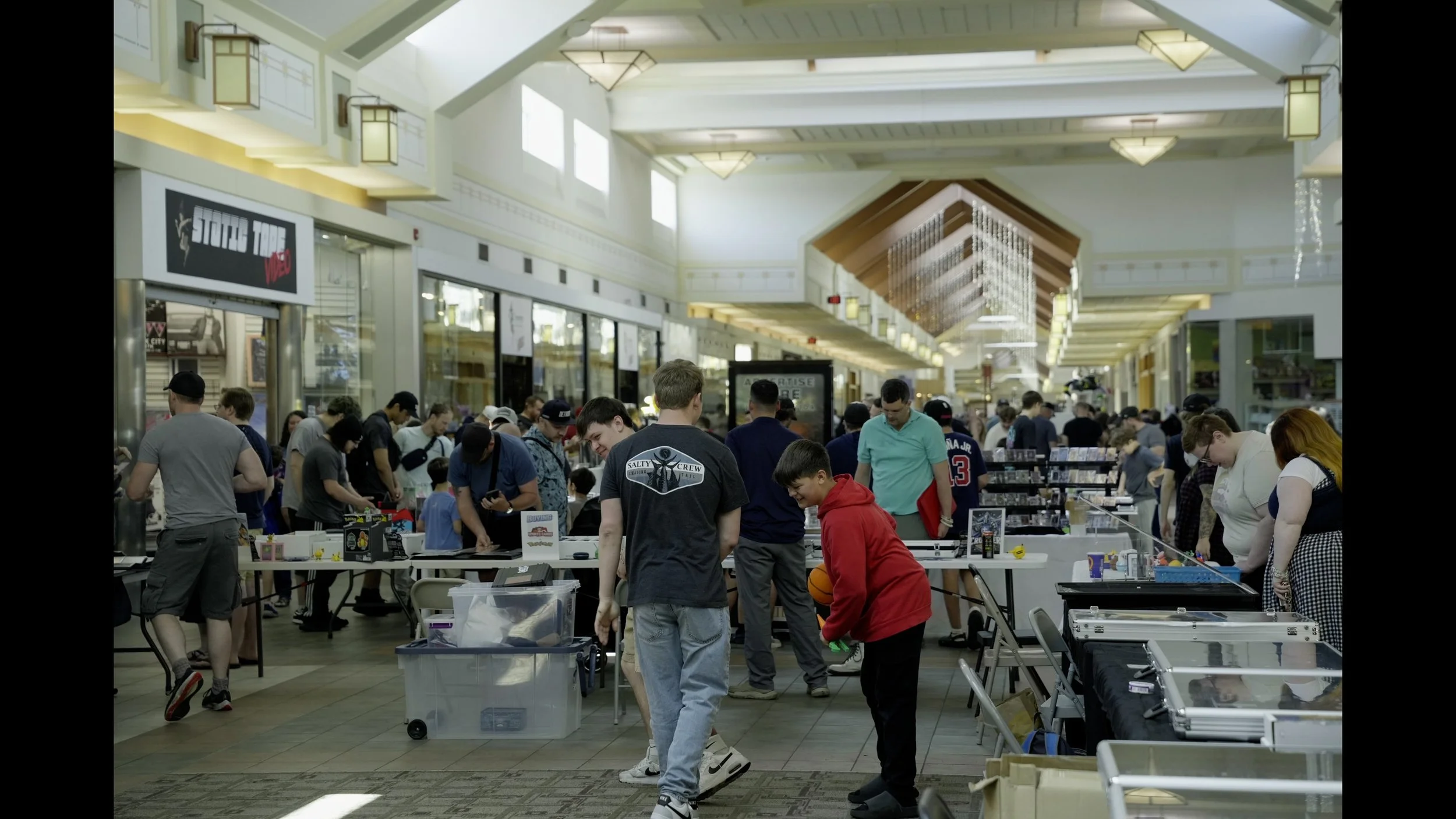 Crowded indoor mall collectibles market with vendors and shoppers browsing trading cards and memorabilia at tables.