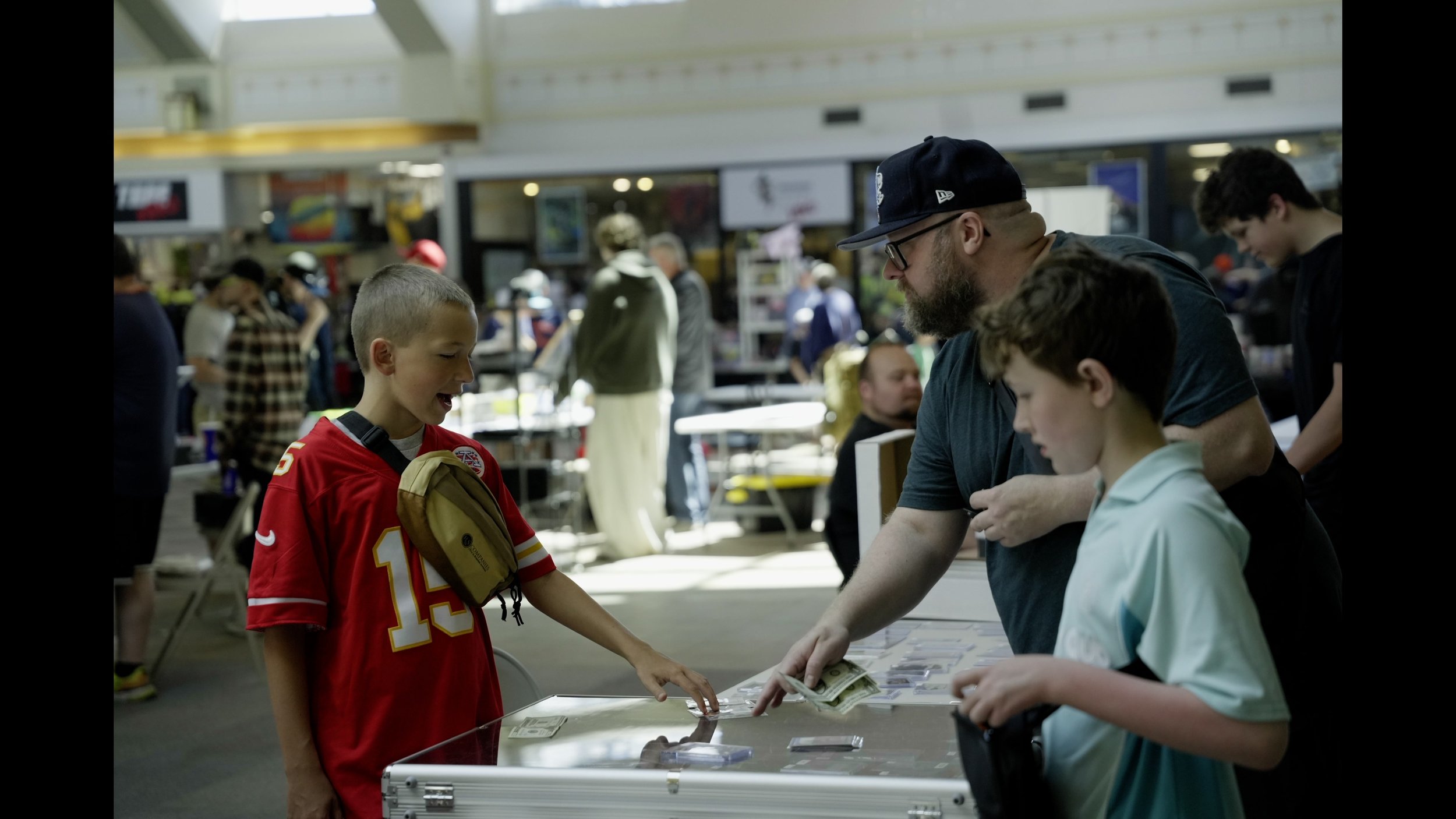 Father and two boys browse sports trading cards at a convention table, with cash and card displays visible.