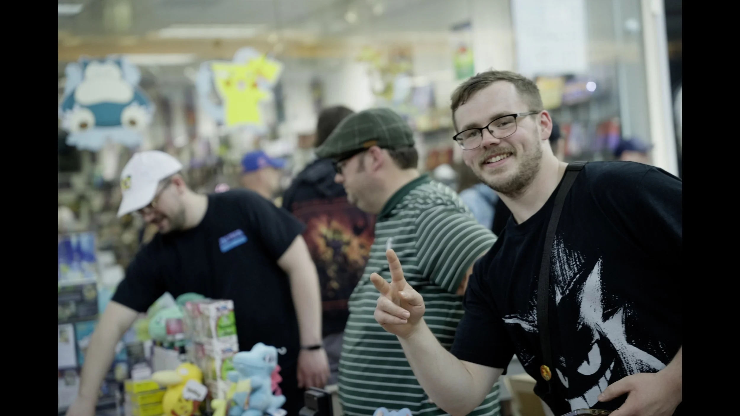 Smiling man with glasses and a black graphic T-shirt poses with friends inside a busy shop.