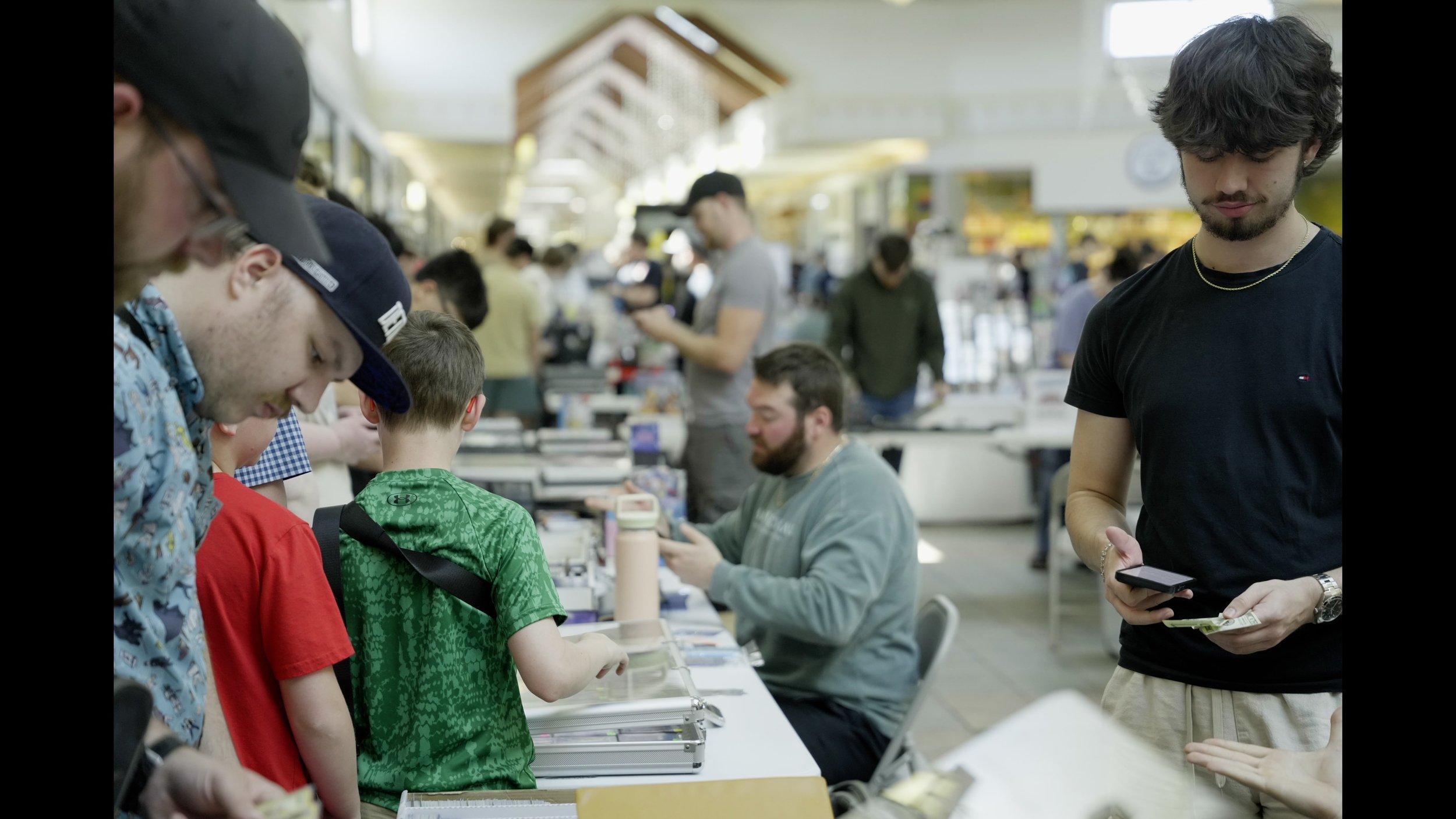 Crowded indoor event with adults and children browsing tables, while a young man checks his phone and holds tickets.
