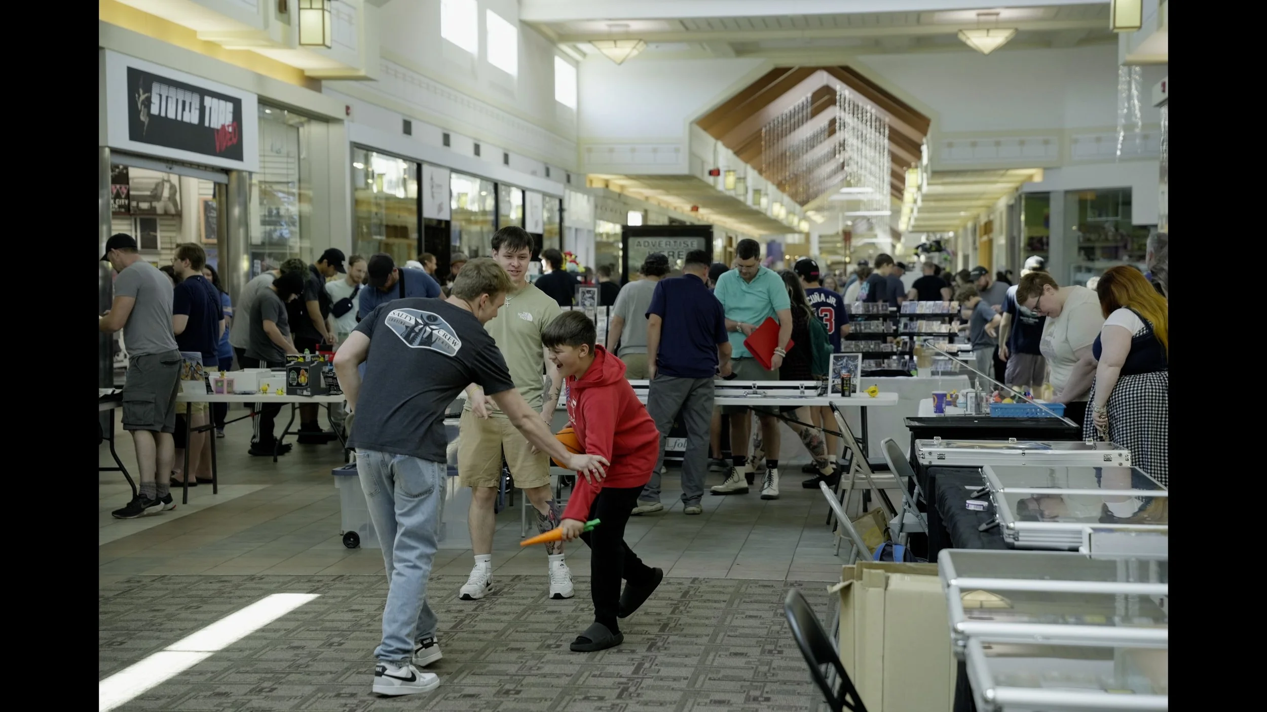 Crowded indoor trading card show with vendors and visitors browsing display tables in a shopping mall.