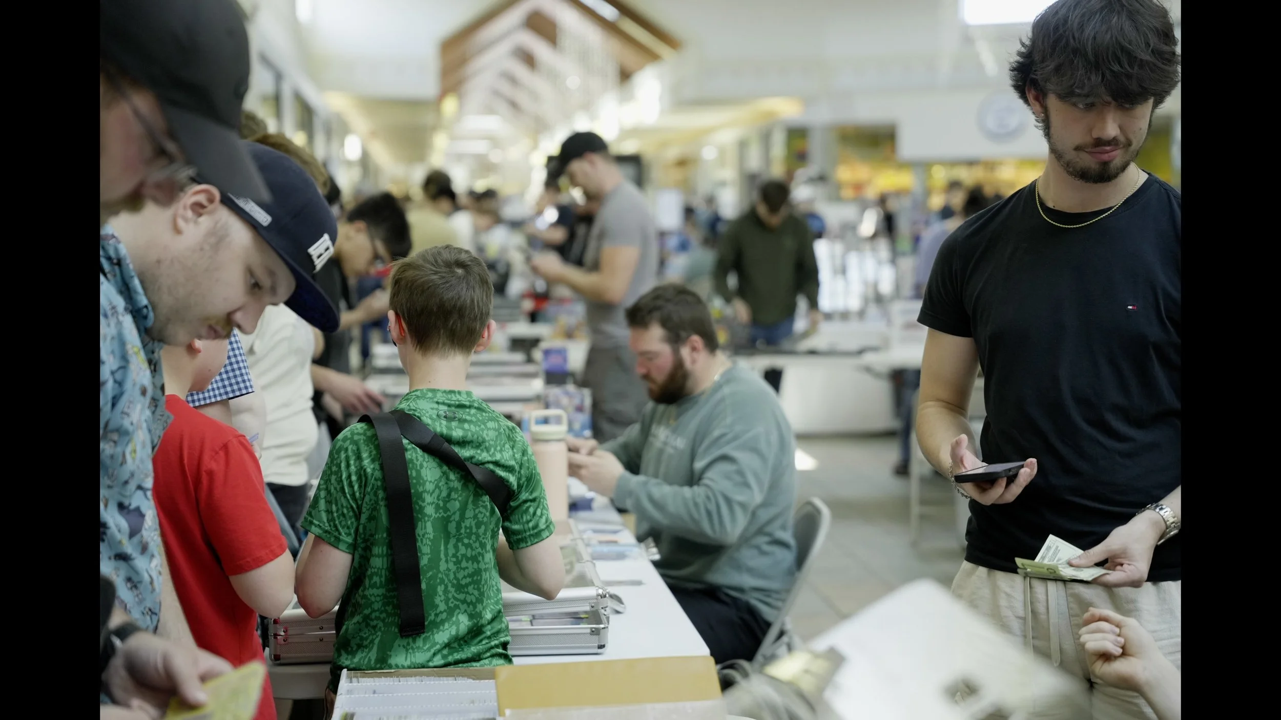 Crowded indoor event with children and adults gathered around a table while a man holds a smartphone and cash.