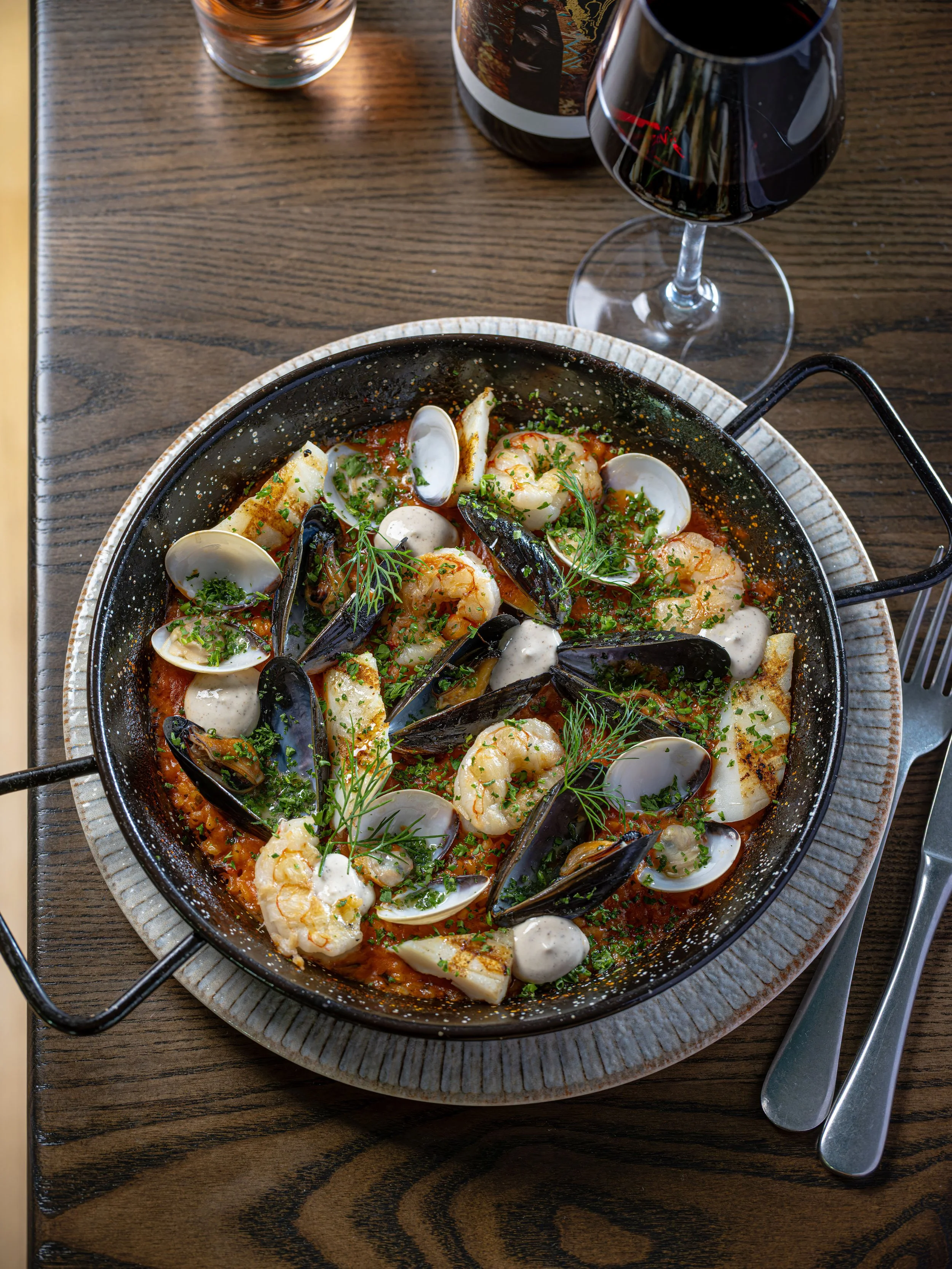 Seafood skillet with shrimp, mussels, clams, and white fish in tomato sauce, garnished with fresh herbs, beside red wine
