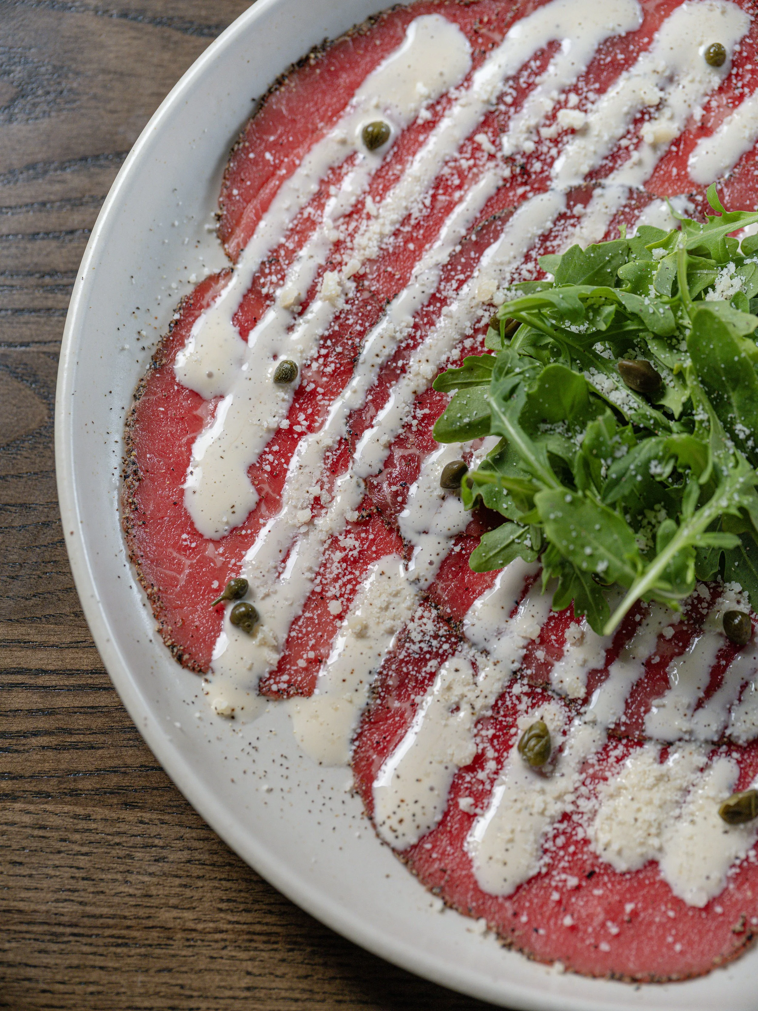 Beef carpaccio topped with creamy sauce, crumbled cheese, capers, and arugula on a white plate