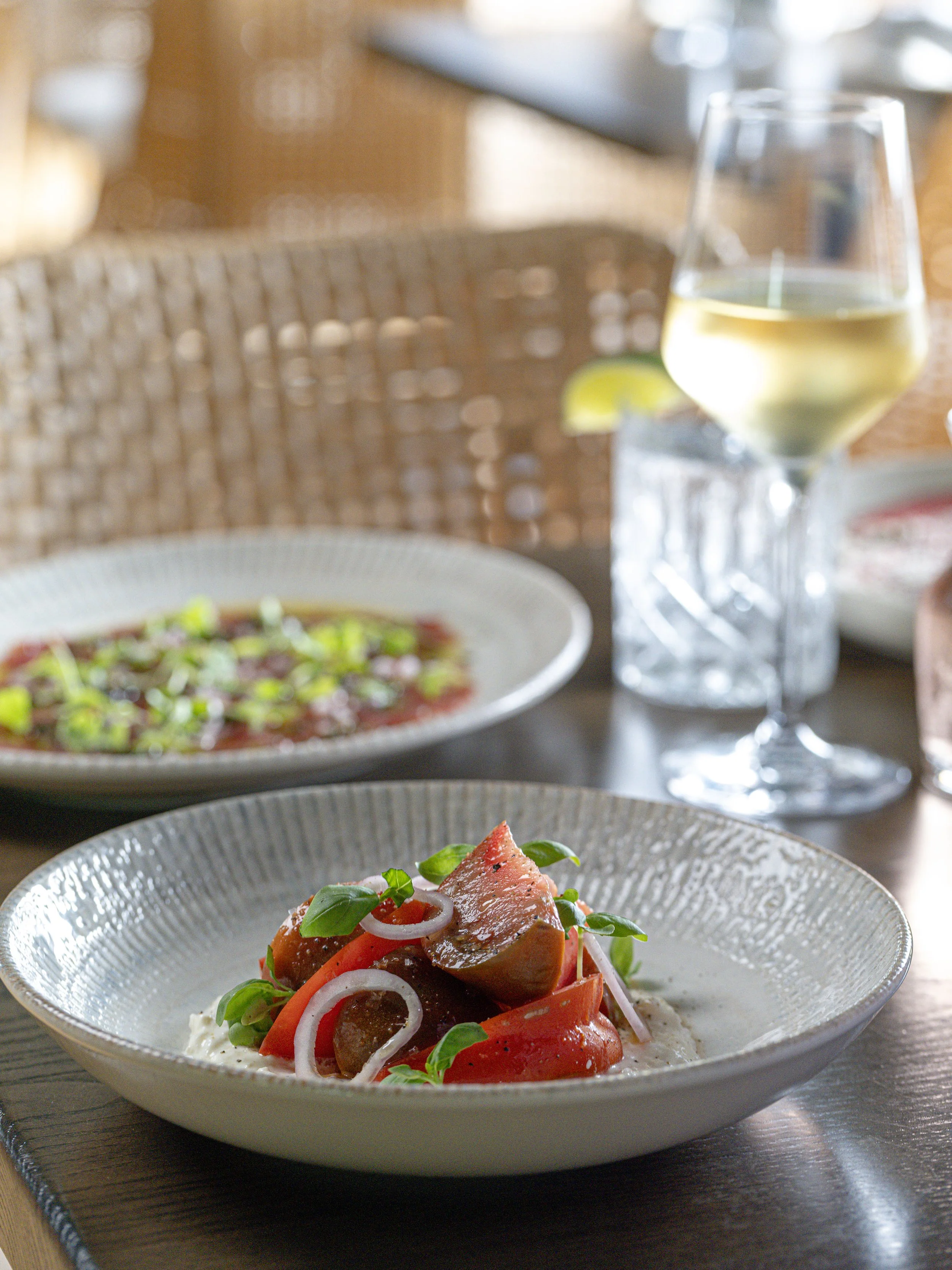 Restaurant table with a plated tomato and grilled meat salad, garnished with greens and onions, alongside a glass of white wine.