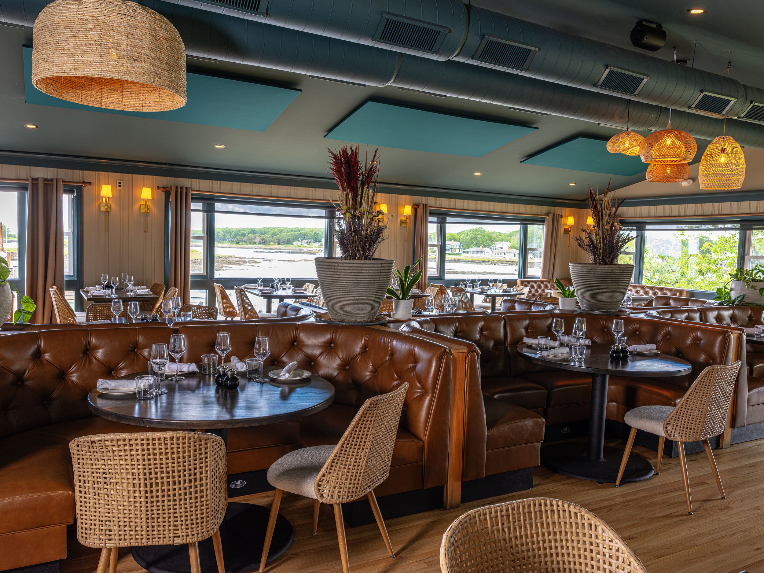 Upscale waterfront restaurant dining room with leather booths, wicker chairs, set tables, pendant lights, and large windows overlooking the shoreline