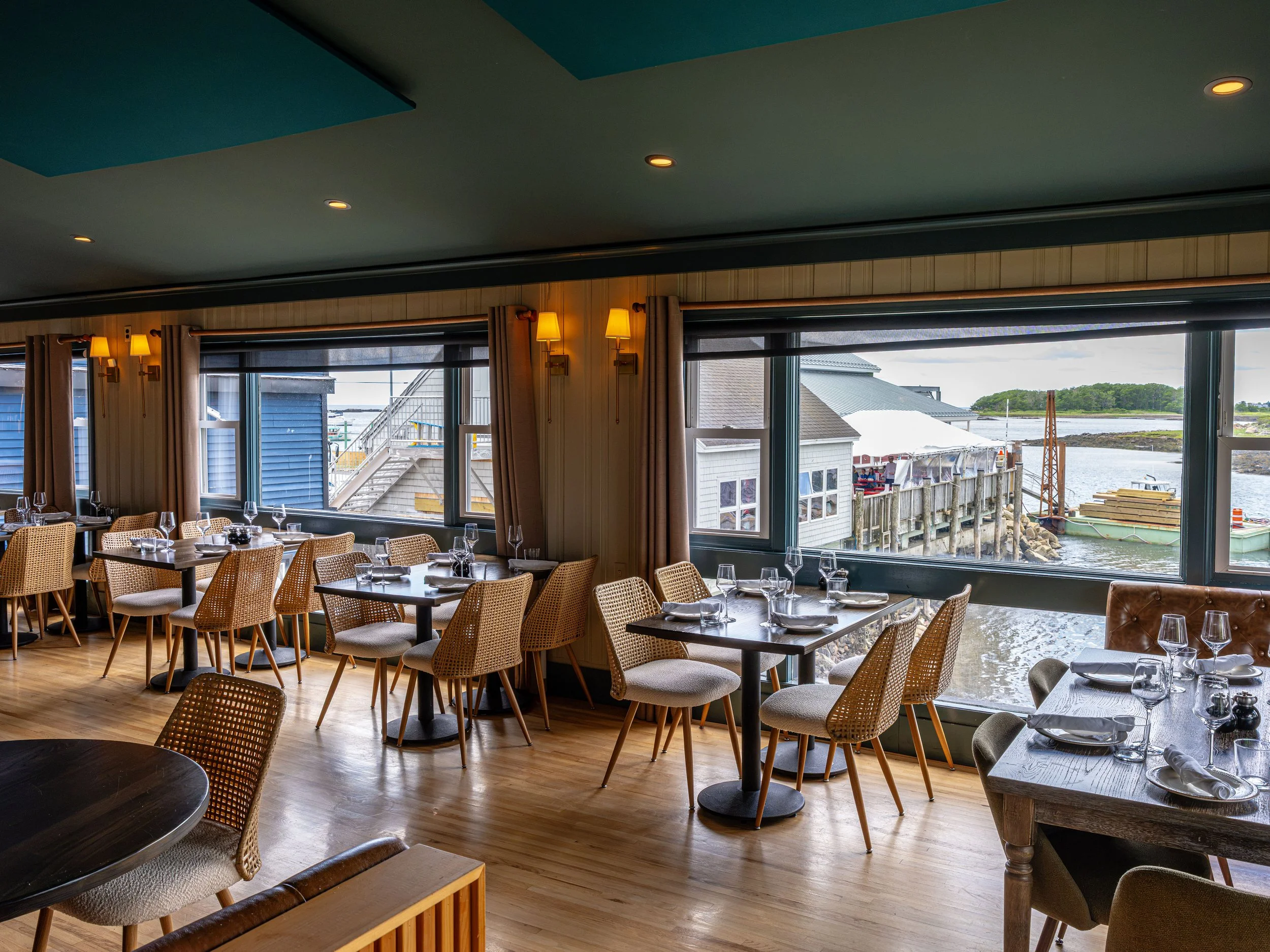 Waterfront restaurant dining room with set tables, woven chairs, large windows, and harbor views