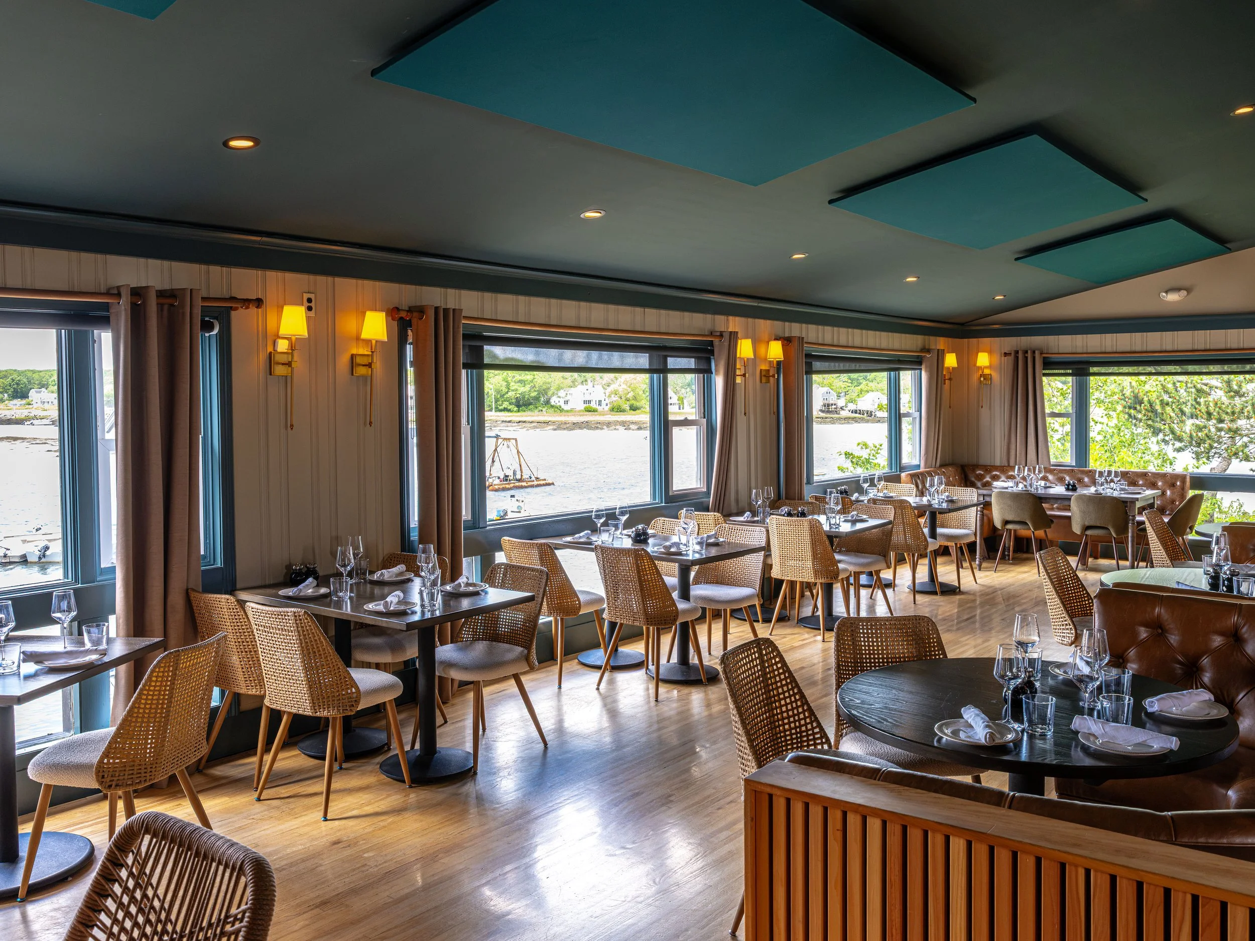 Elegant waterfront restaurant dining room with set tables, woven chairs, leather booths, and large windows overlooking the water