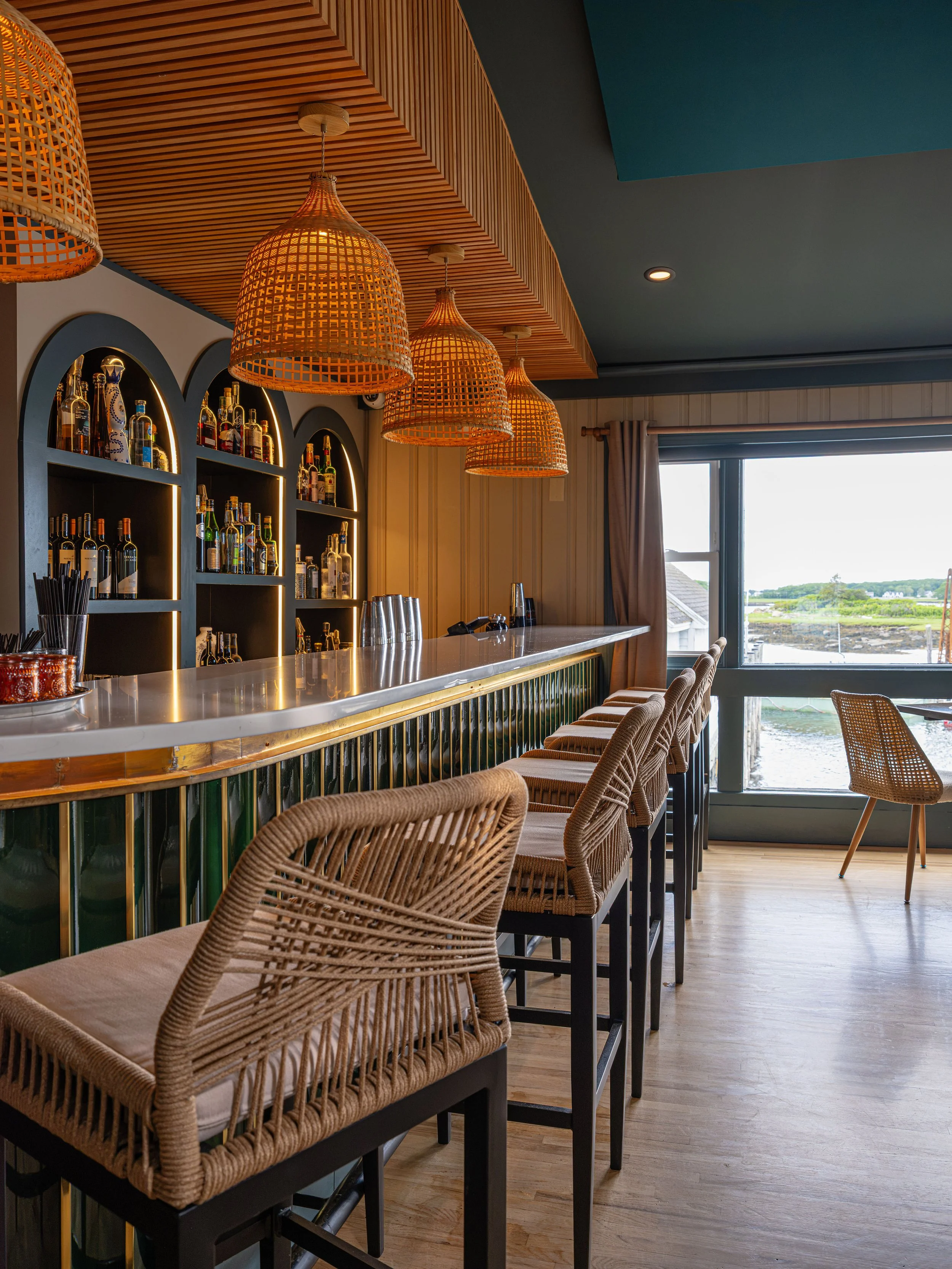 Stylish restaurant bar with woven pendant lights, liquor shelves, and wicker bar stools overlooking a waterfront