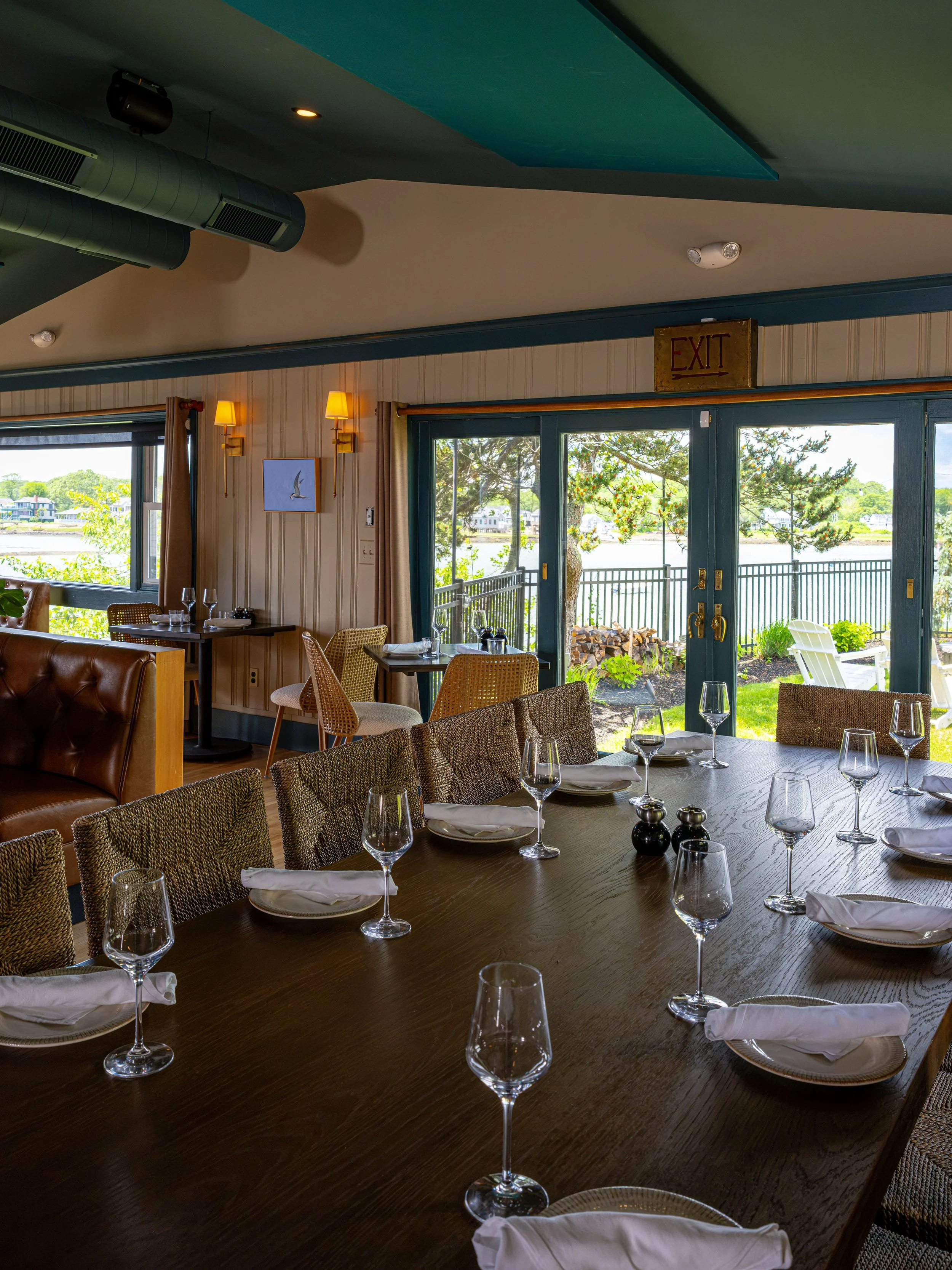 Elegant waterfront restaurant dining room with set tables, wine glasses, woven chairs, and large windows overlooking the water