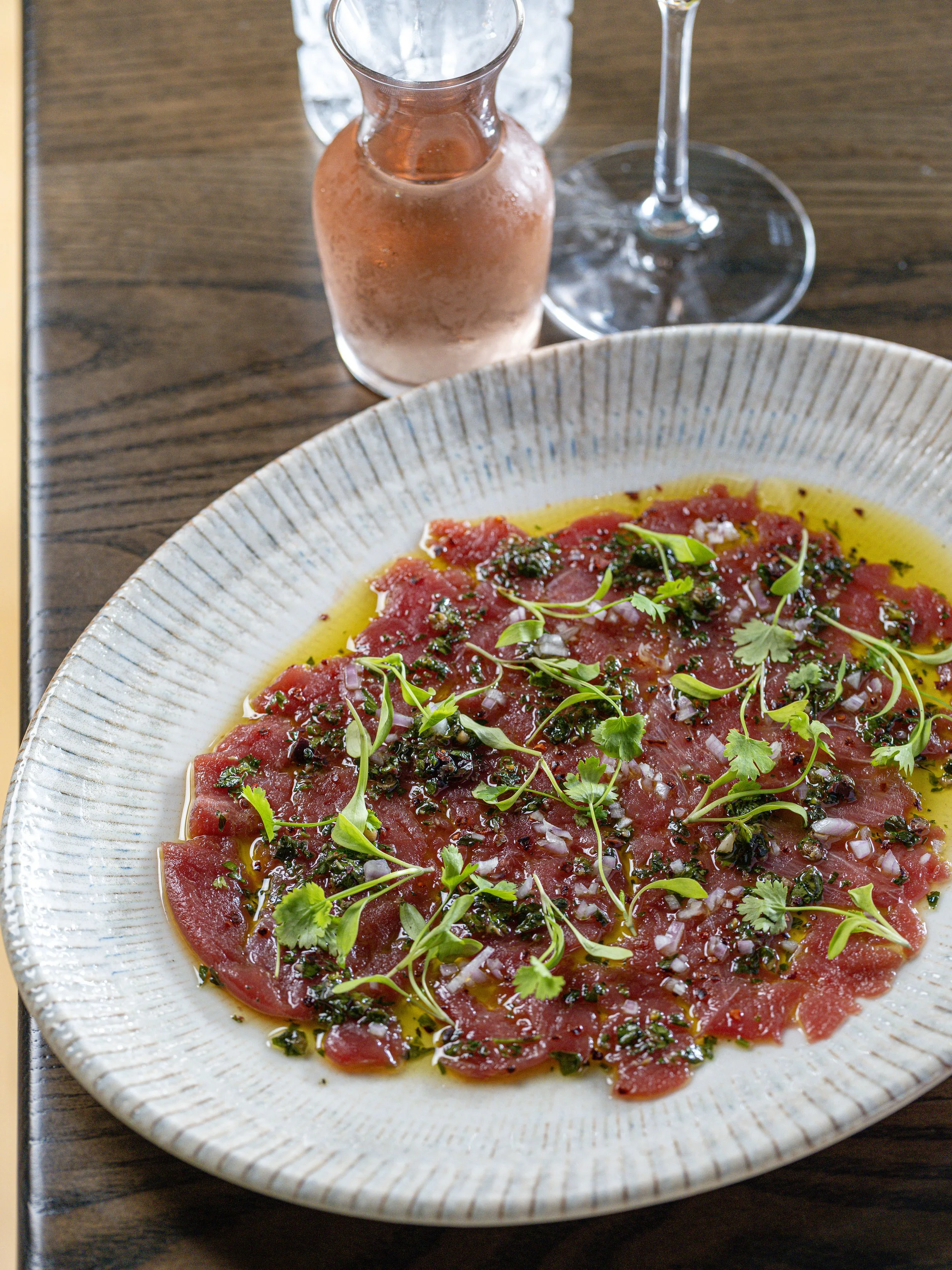 Thinly sliced raw tuna carpaccio topped with herbs, diced onions, and olive oil on a ceramic plate, with a pink drink and wine glass in the background.