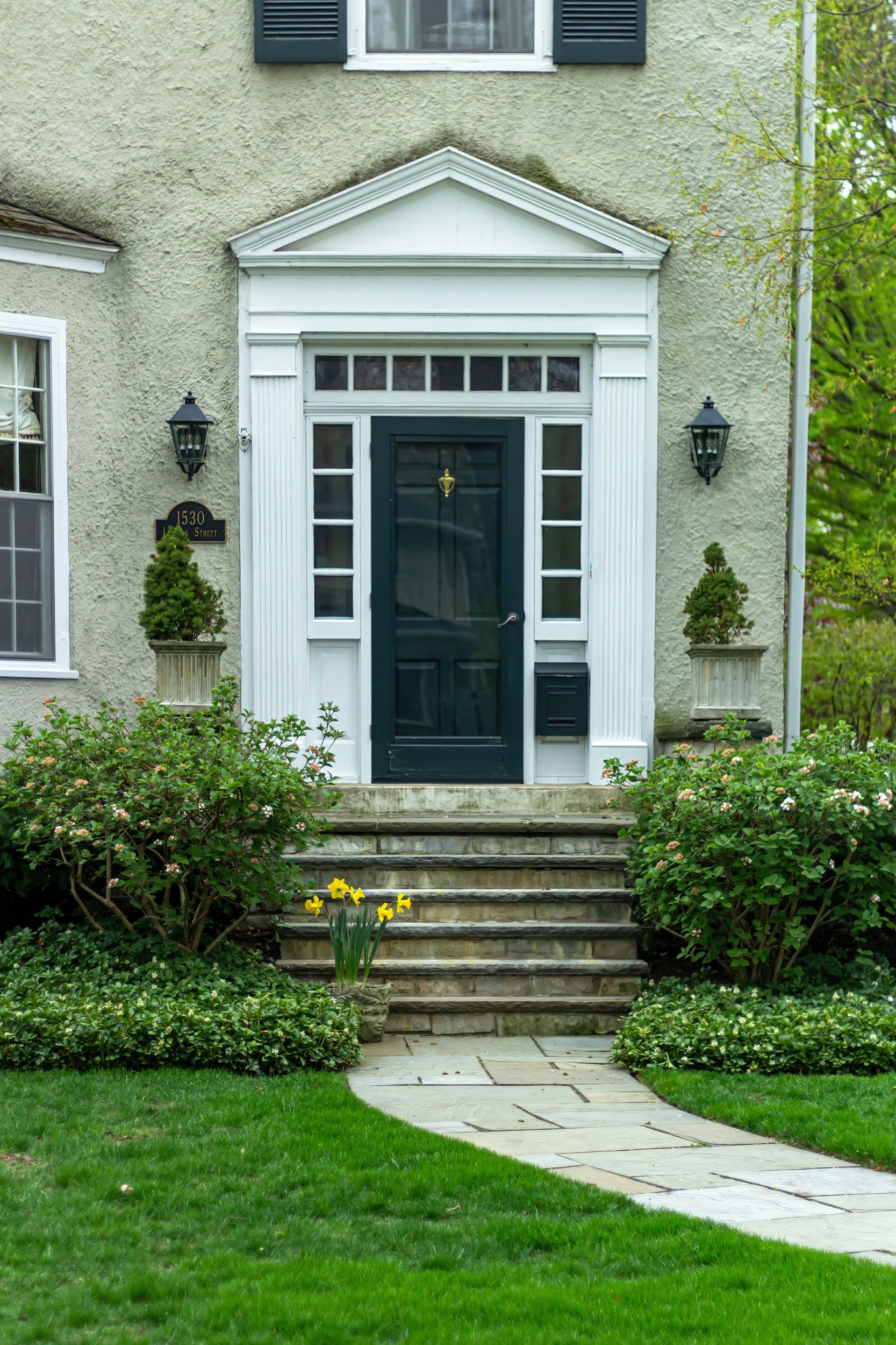Elegant green stucco home entrance with black door, white classical trim, lanterns, stone steps, manicured shrubs, and a walkway