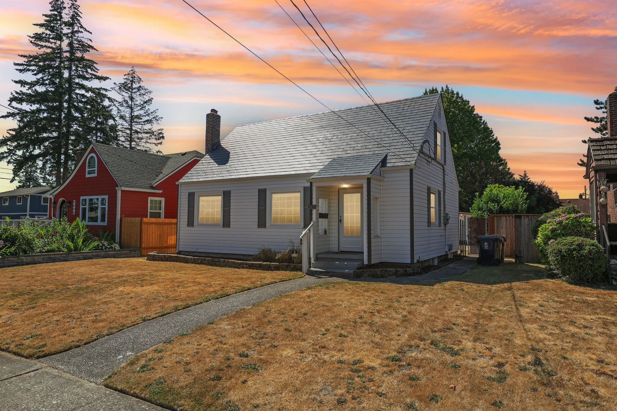 Residential neighborhood with a white house, red home, dry lawns, trees, and colorful sunset sky