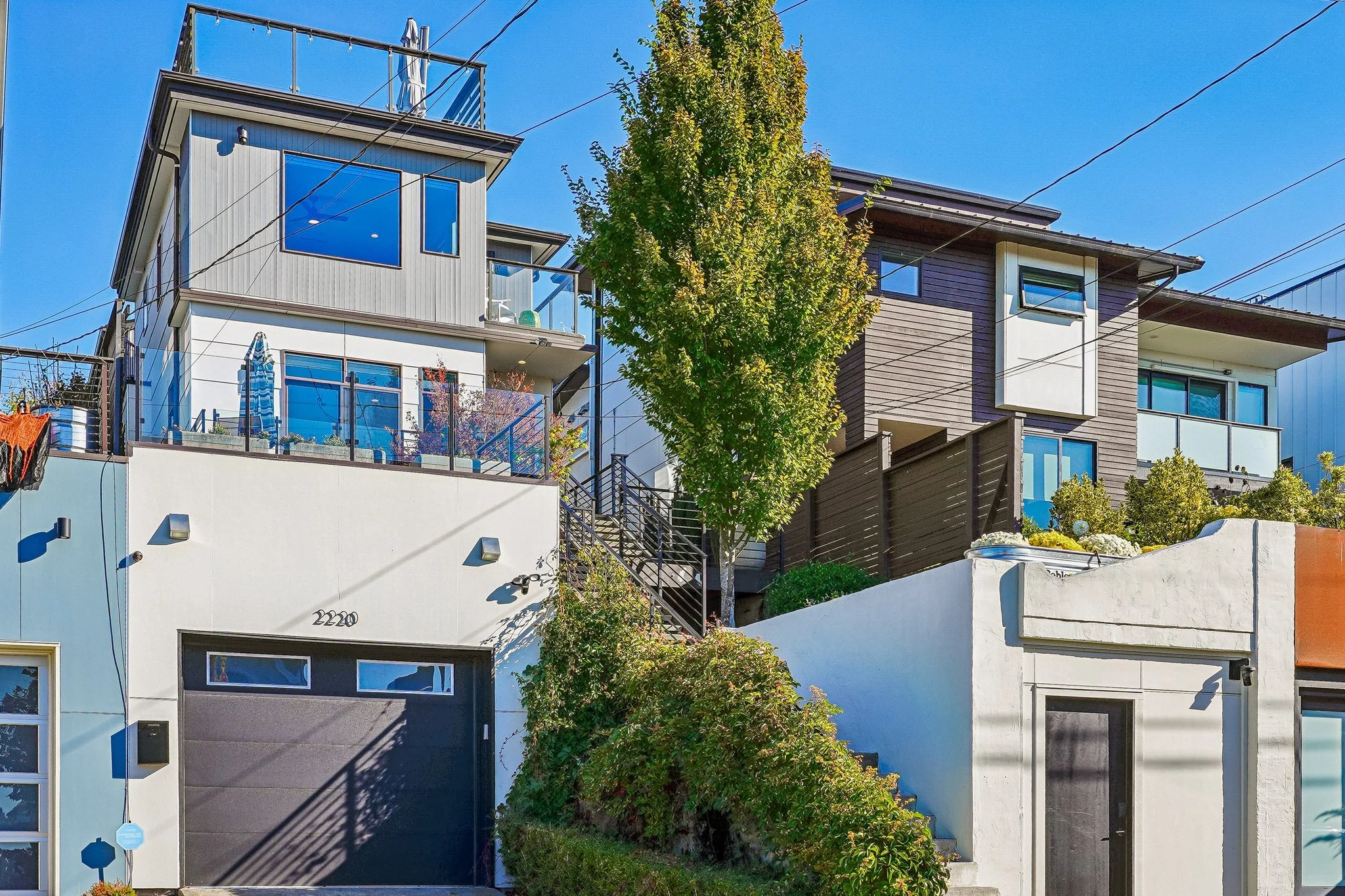 Modern urban townhouses with balconies, landscaped front gardens, and overhead utility wires under a clear blue sky