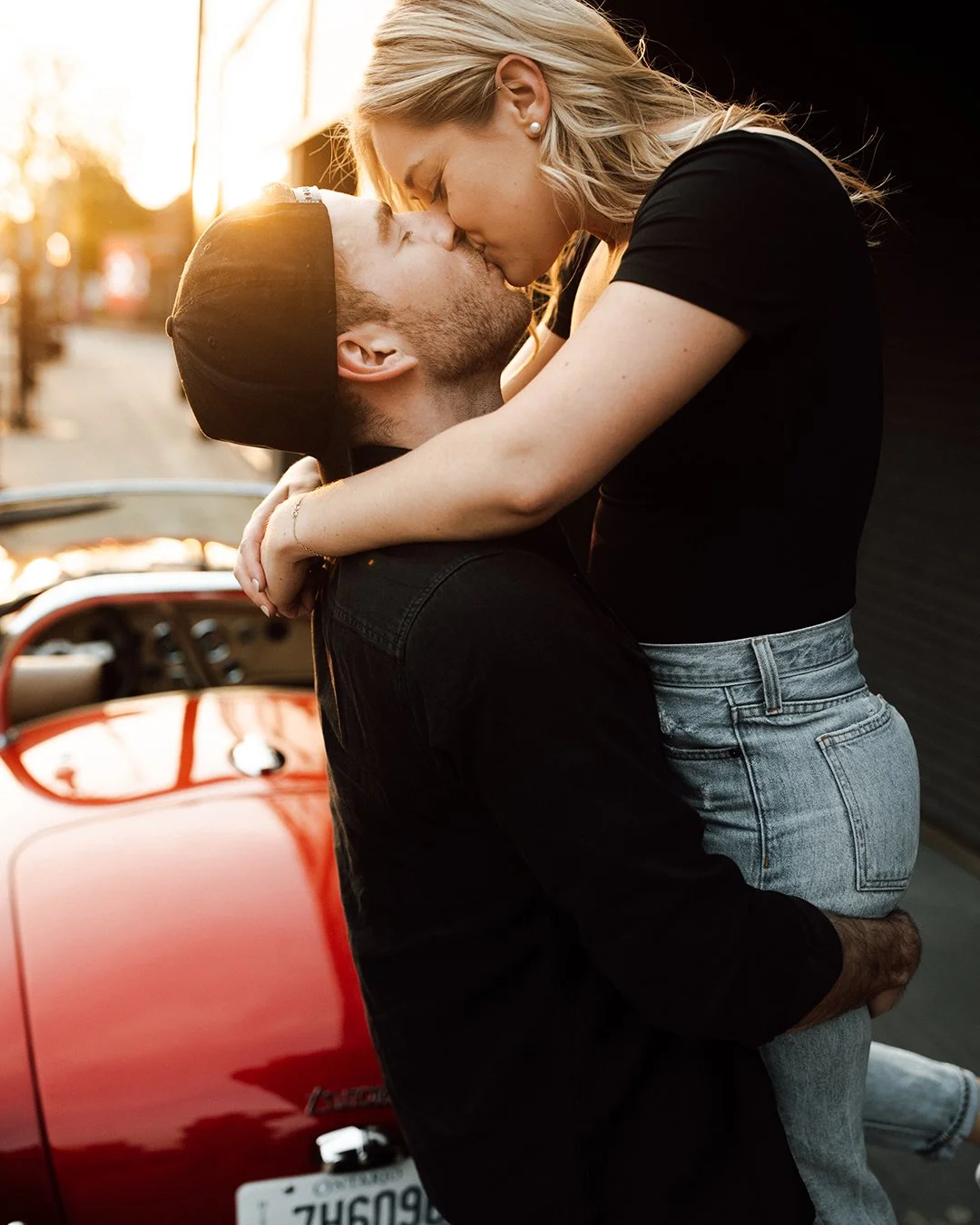 man hoisting a woman backlit by sun and a red sportscar
