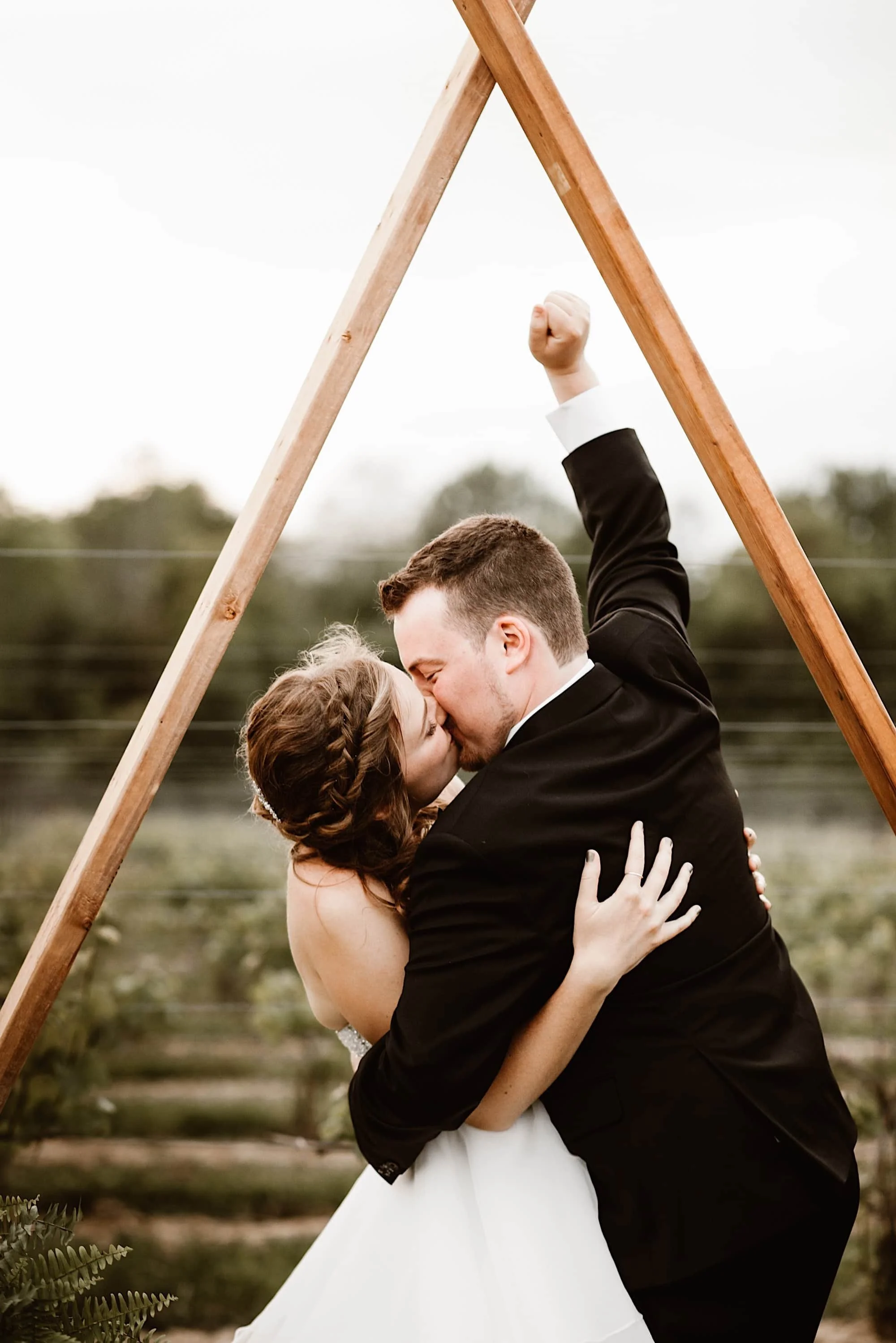 groom raises fist under triangular archway as he kisses his bride 