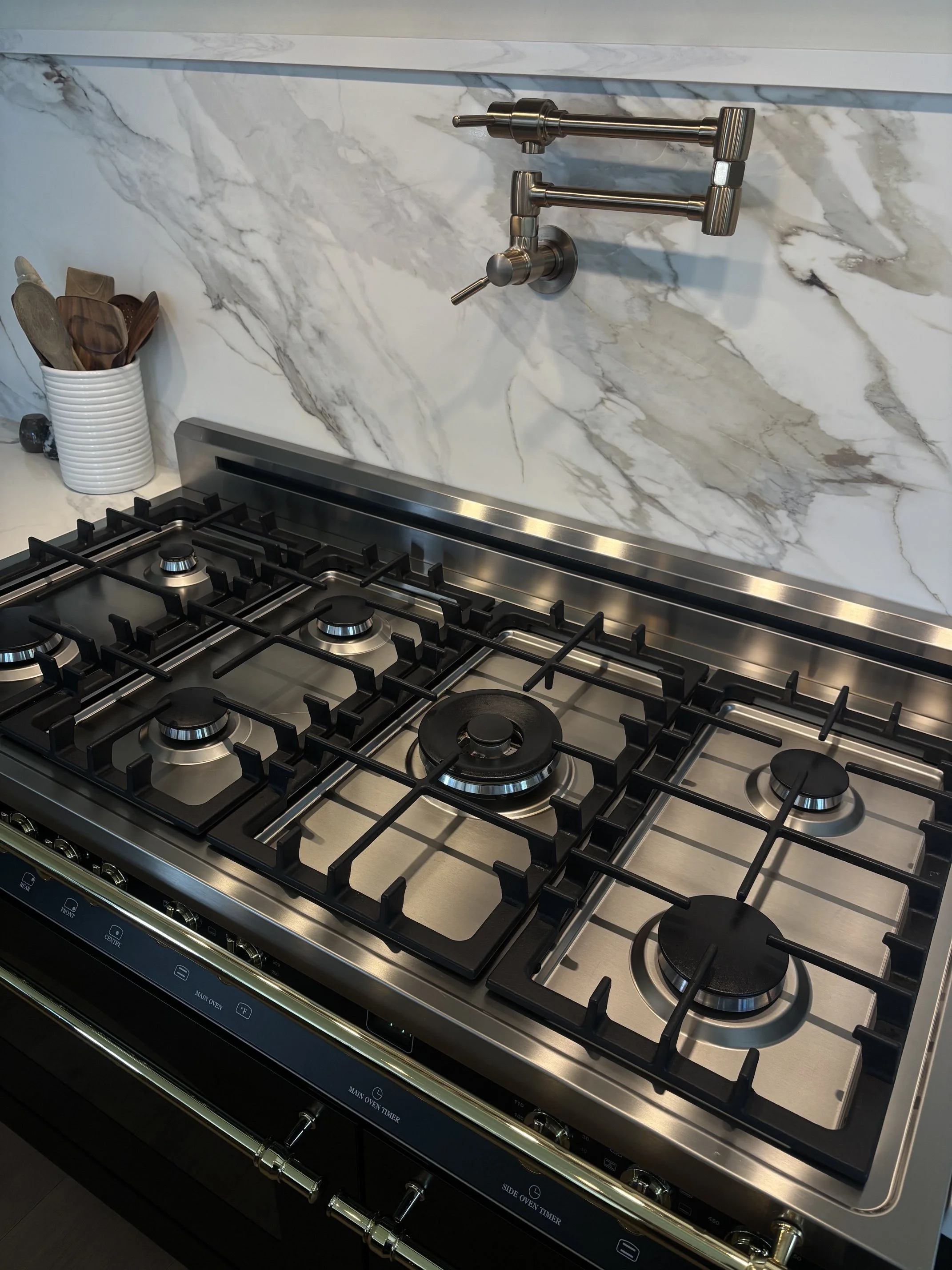 Modern stainless steel gas range with multiple burners, brass handles, wall-mounted pot filler, and marble backsplash