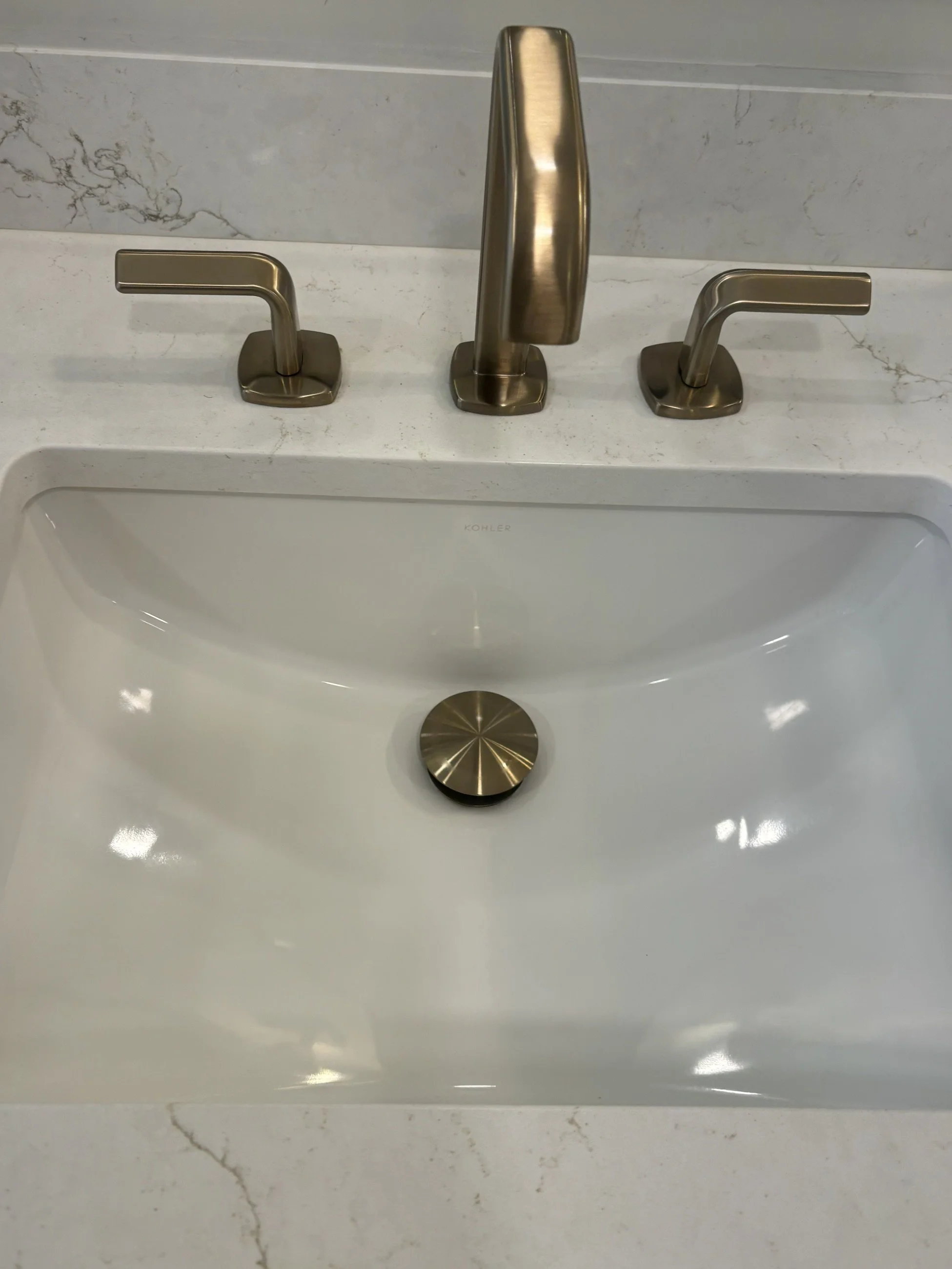 Brushed bronze three-hole bathroom faucet with two lever handles and matching drain on a white Kohler sink