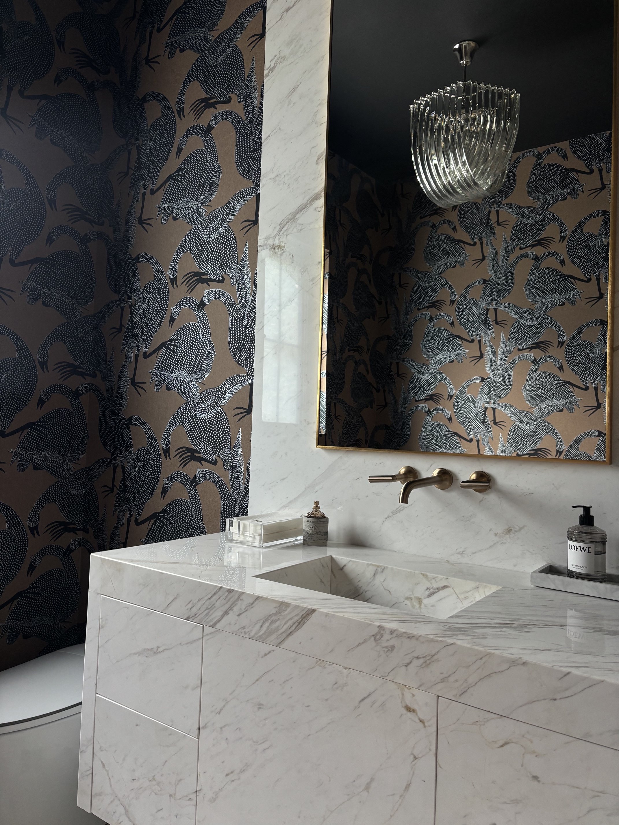 Luxury bathroom with white marble vanity, patterned bird wallpaper, brass faucet, mirror, and glass pendant light