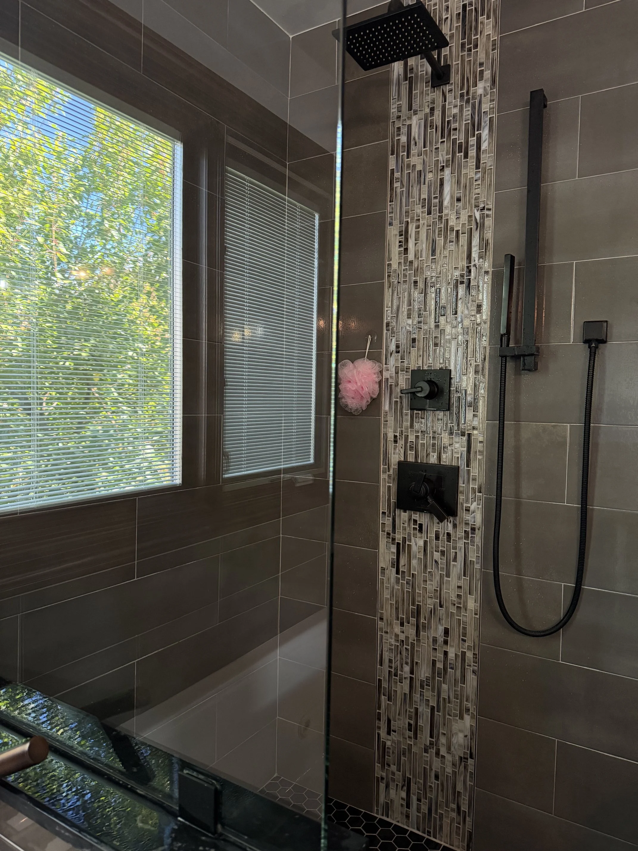 Modern tiled bathroom shower with black fixtures, handheld showerhead, mosaic accent wall, window blinds, and pink bath sponge.