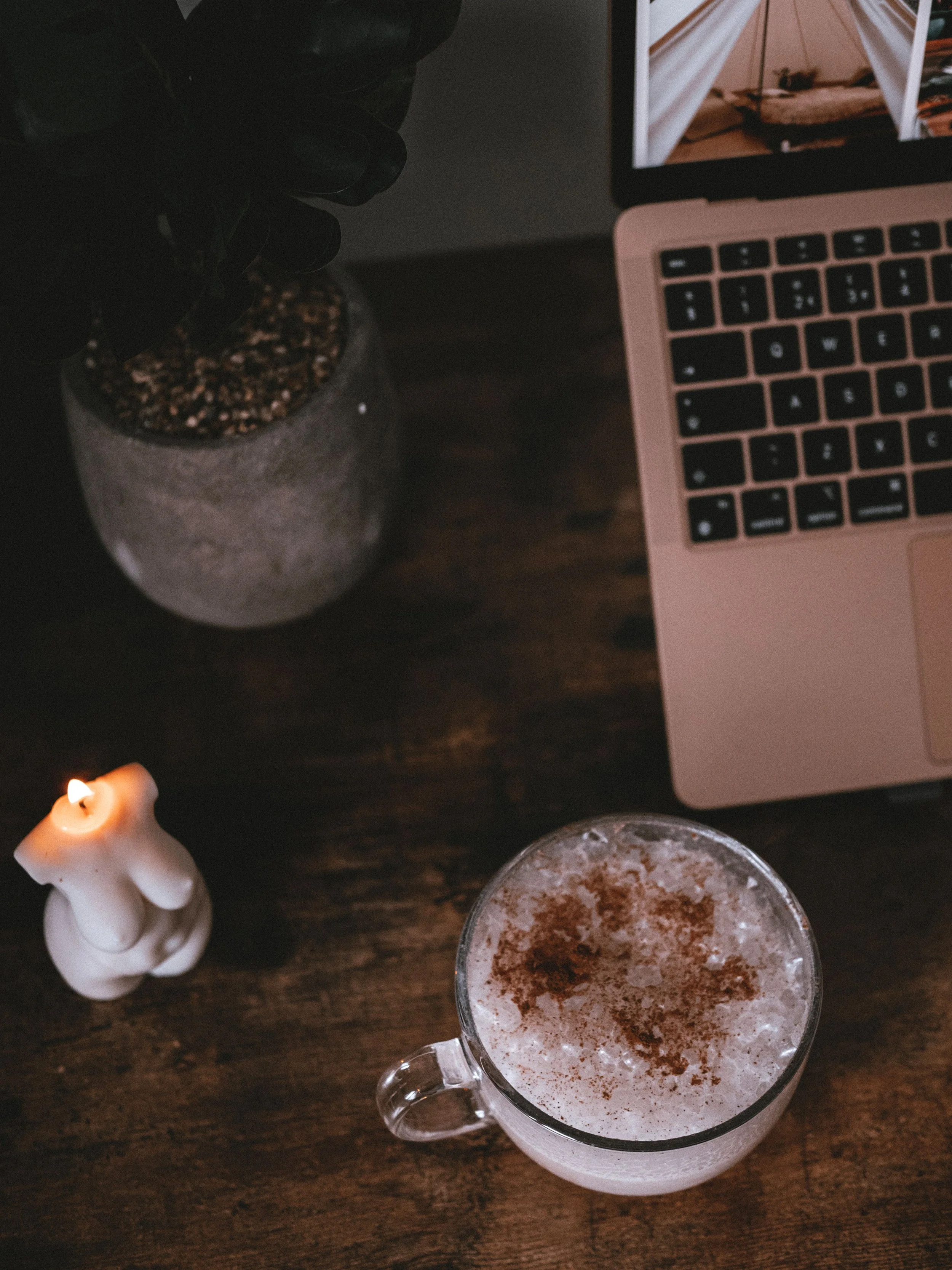 Cozy workspace with a pink laptop, potted plant, lit candle, framed photo, and frothy cocoa-dusted drink on a wooden desk