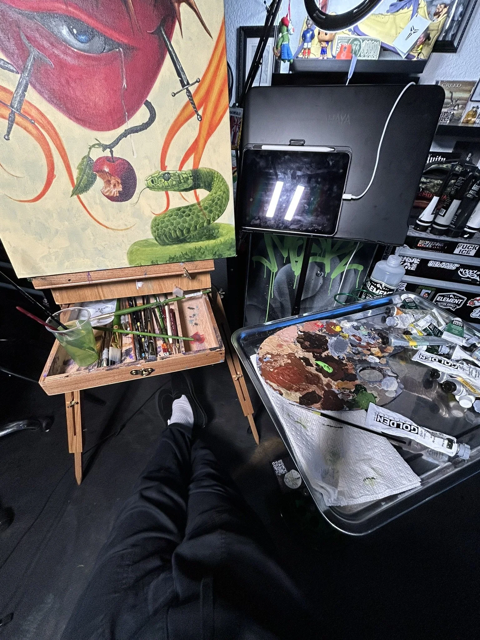 Artist's workspace with a painting of a red-masked figure and green snake, brushes, easel, palette, paint tubes, and art supplies.