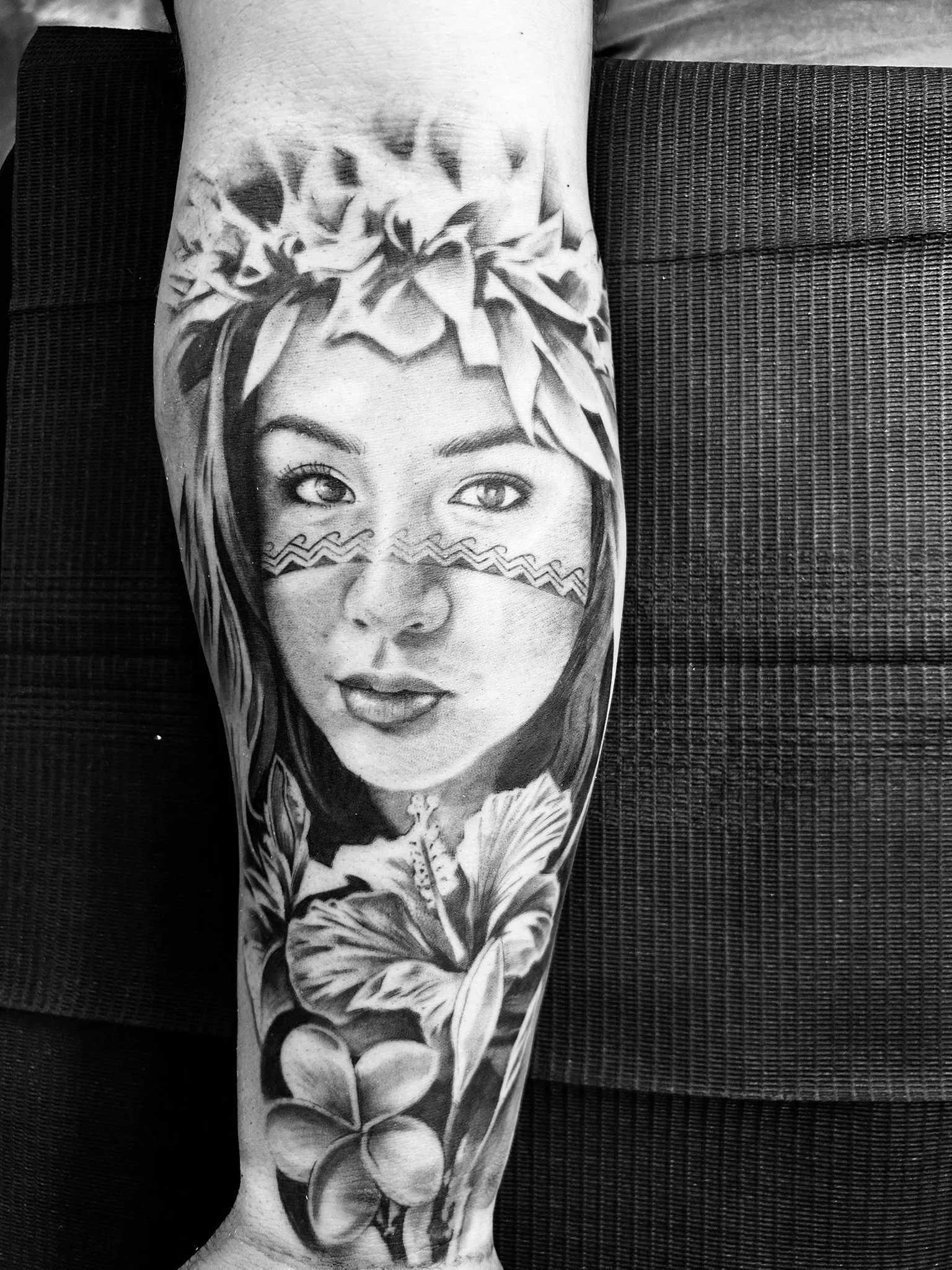 Black-and-white photo of a detailed tattoo featuring a woman’s face with floral elements, leaves, and geometric face markings on a forearm.