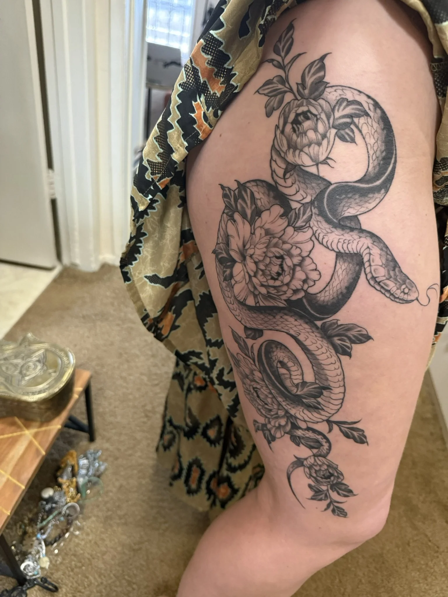 Close-up of an intricate black-and-gray thigh tattoo featuring a coiled dragon surrounded by flowers and leaves.