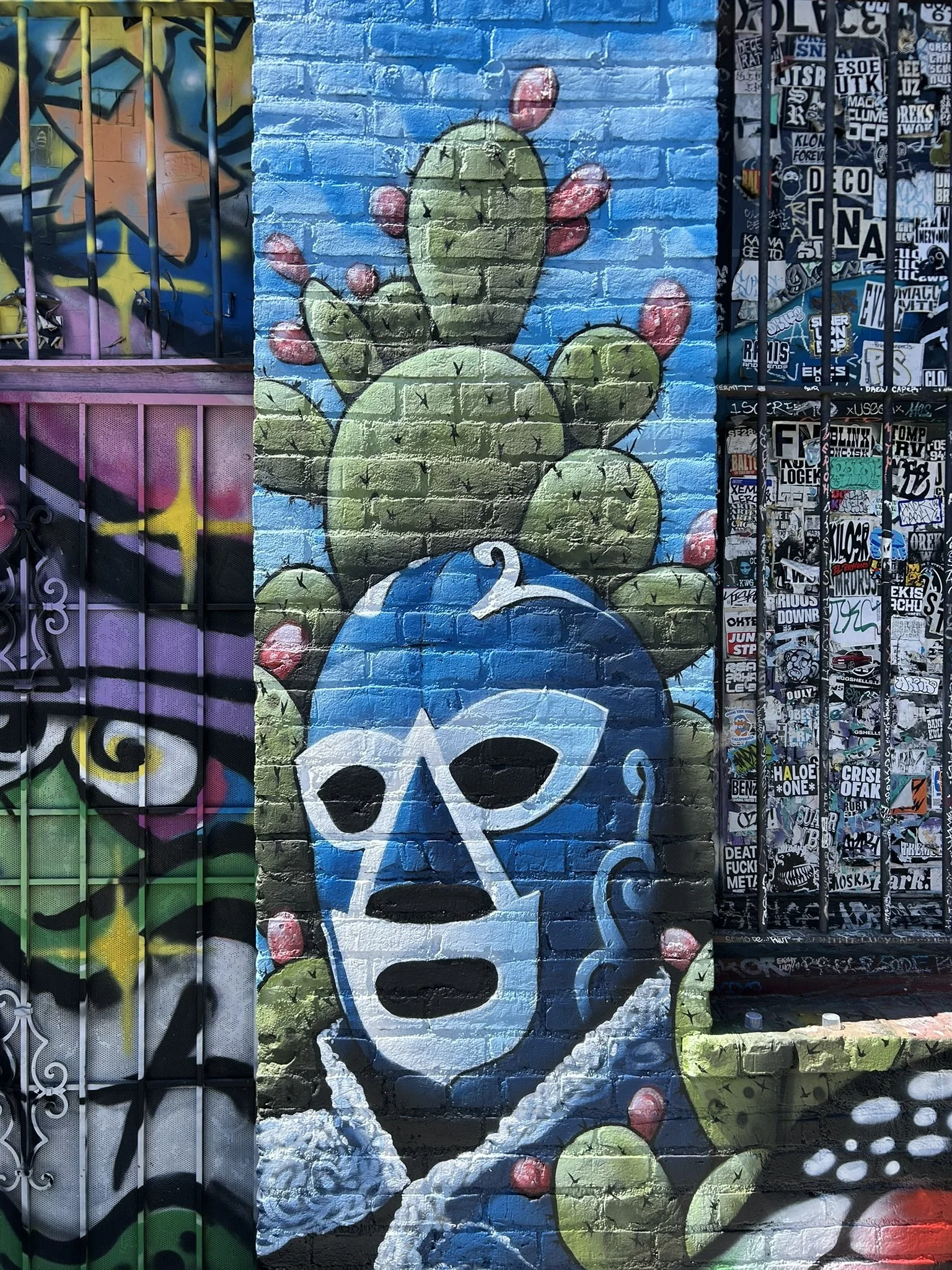 Colorful street art mural of a green cactus character surfing a blue wave on a brick wall, surrounded by graffiti and sticker-covered surfaces.