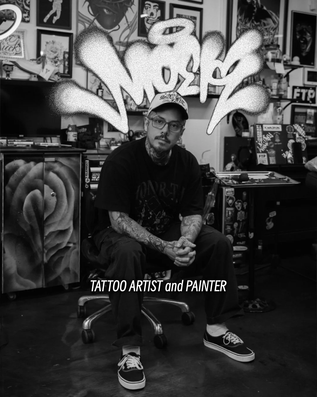 Black-and-white portrait of a tattoo artist and painter seated in a tattoo studio surrounded by artwork.