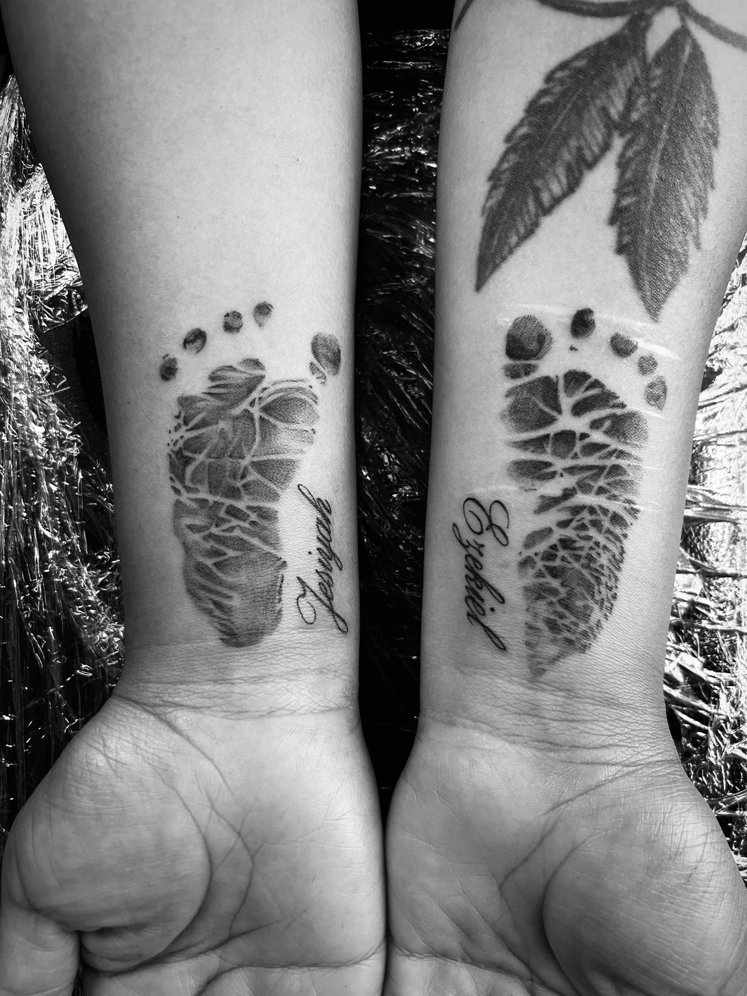Black-and-white photo of two tattooed forearms featuring botanical designs, leaves, and cursive lettering.