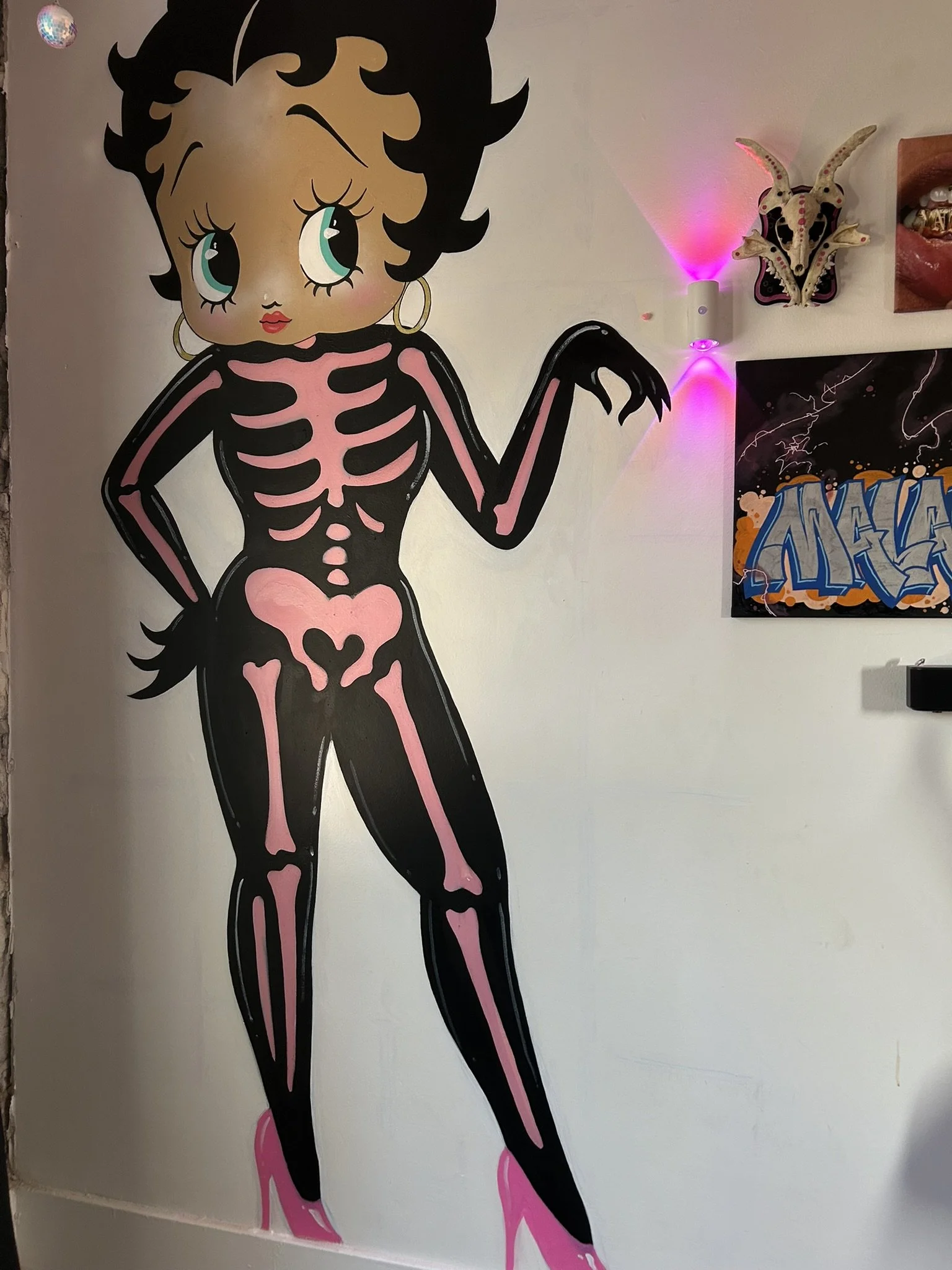 Large Betty Boop-inspired skeleton mural in a black and pink outfit, surrounded by eclectic wall art and pink-purple lighting