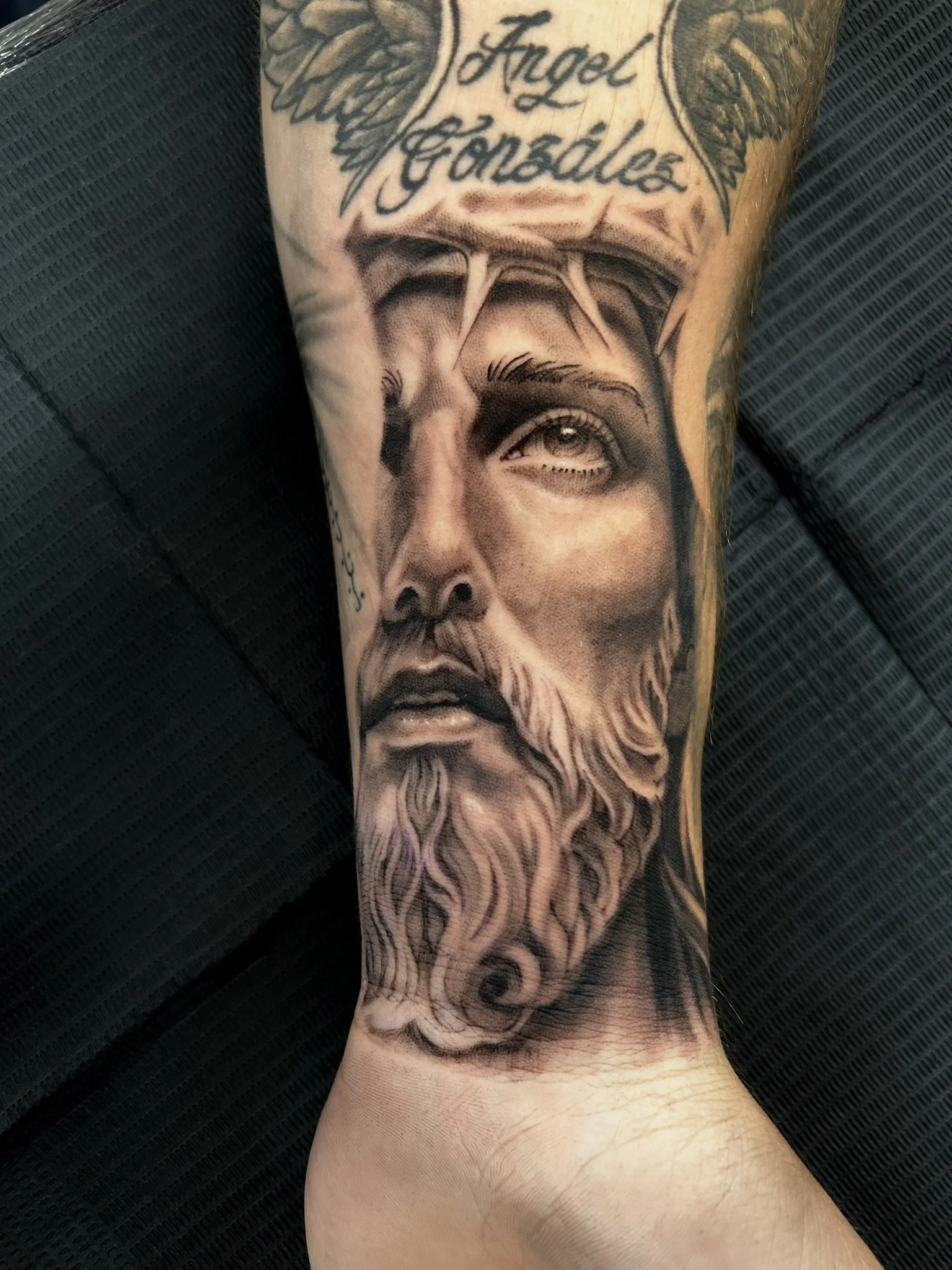Black-and-gray tattoo of a bearded man's portrait with angel wings and cursive lettering on a lower leg