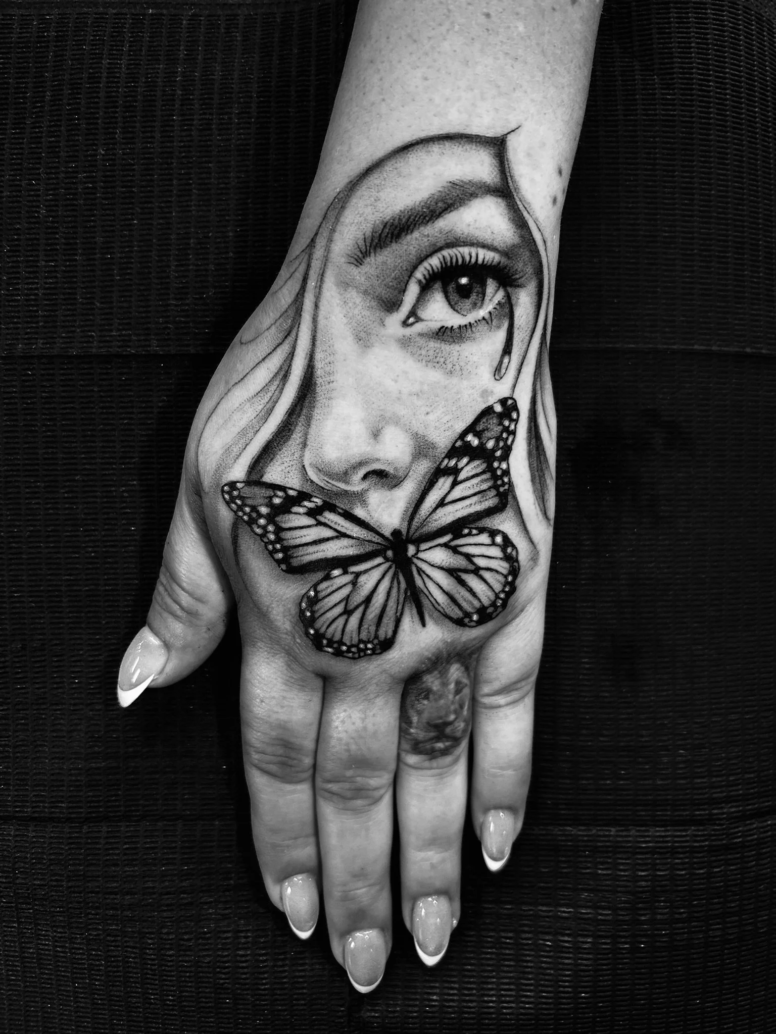 Black-and-white hand tattoo featuring a realistic eye with a tear and butterfly covering the mouth.