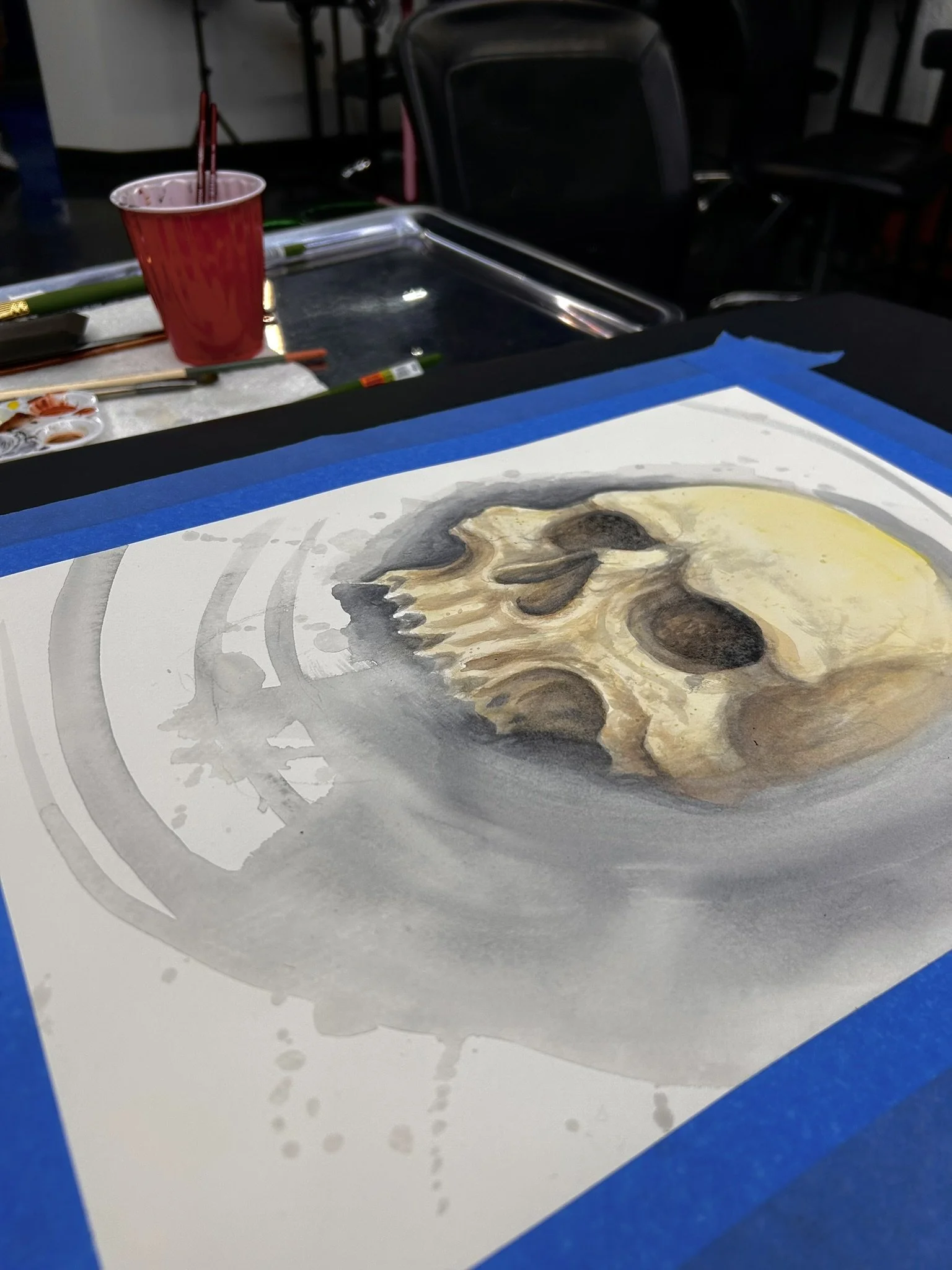 Watercolor painting of a skull on paper taped to a table, with brushes, paint palettes, and a red paint cup nearby