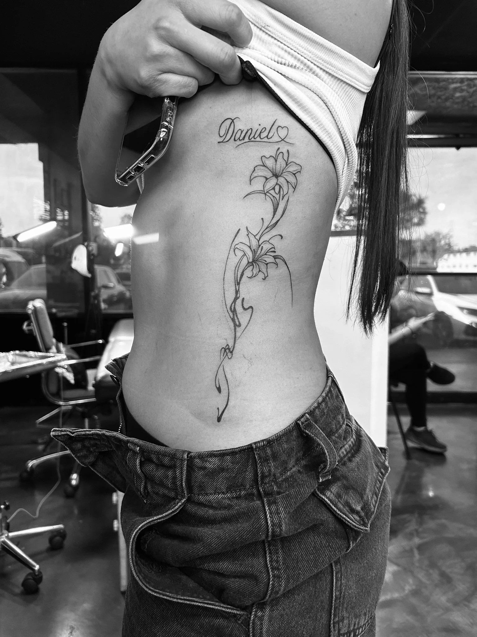 Black-and-white close-up of a woman's side torso showing a vertical floral tattoo with the name “Daniel” and a heart.