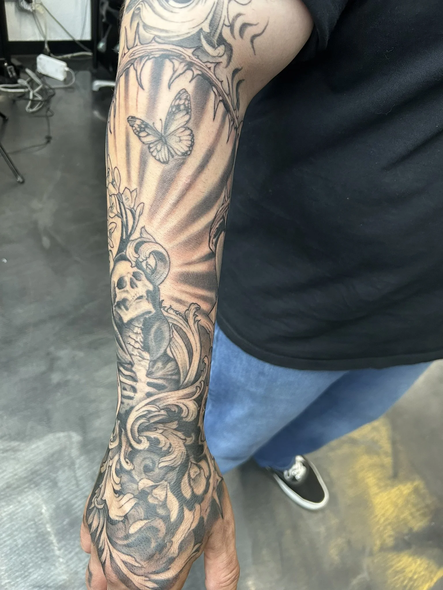 Close-up of a person's arm covered in intricate black-and-gray tattoos, including a butterfly, skull, flowers, and ornamental patterns.