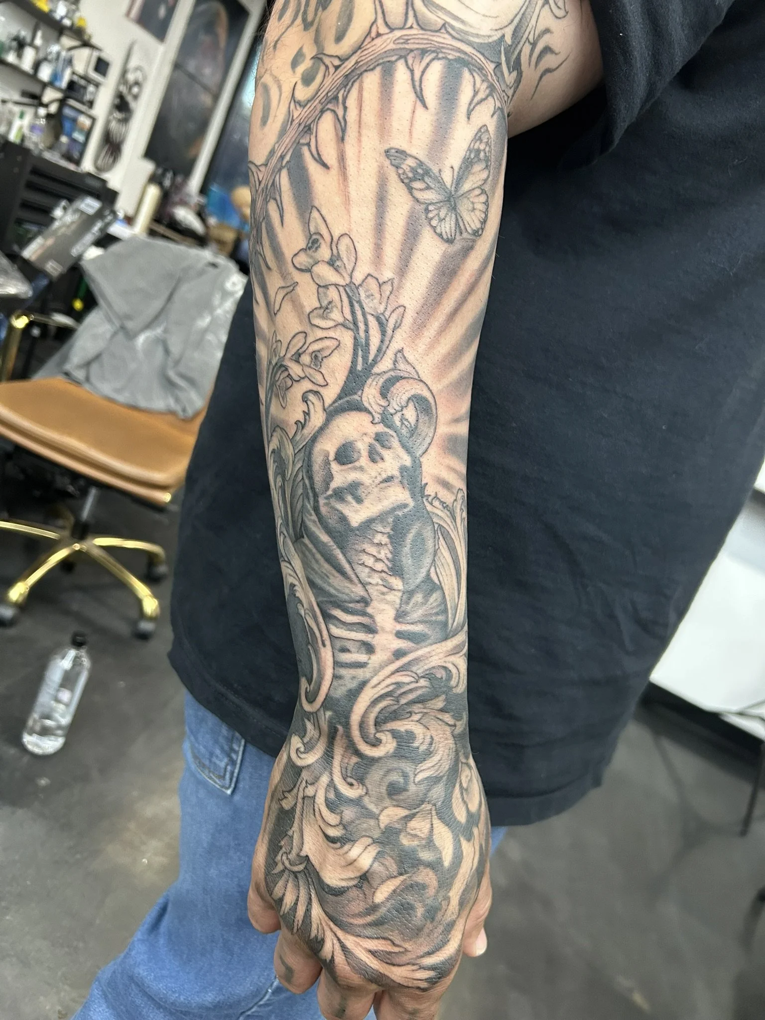 Close-up of a detailed black-and-gray tattoo sleeve featuring a skull, butterfly, flowers, leaves, and ornate patterns on a forearm and hand.