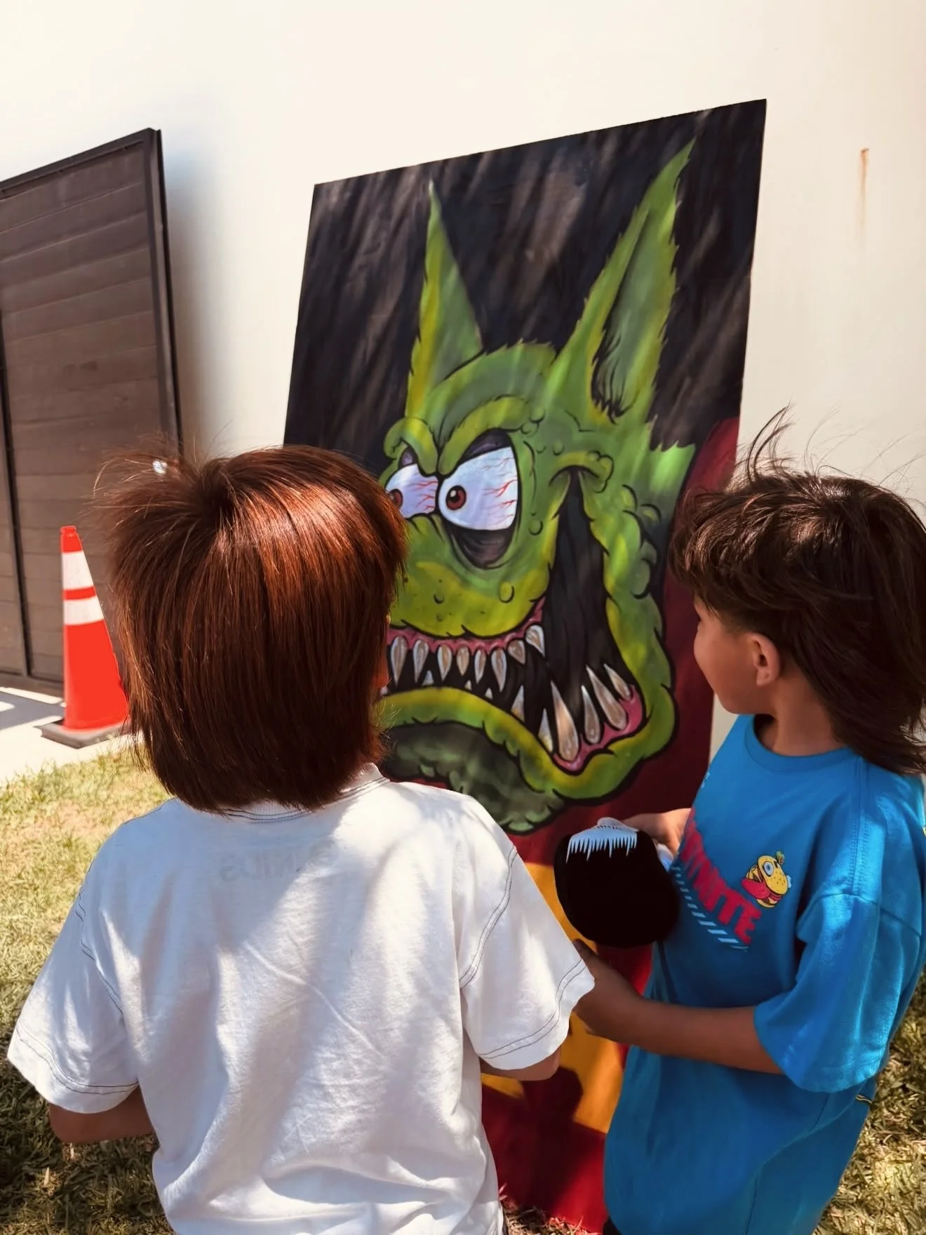 Children play a carnival game in front of a large green monster painting outdoors.